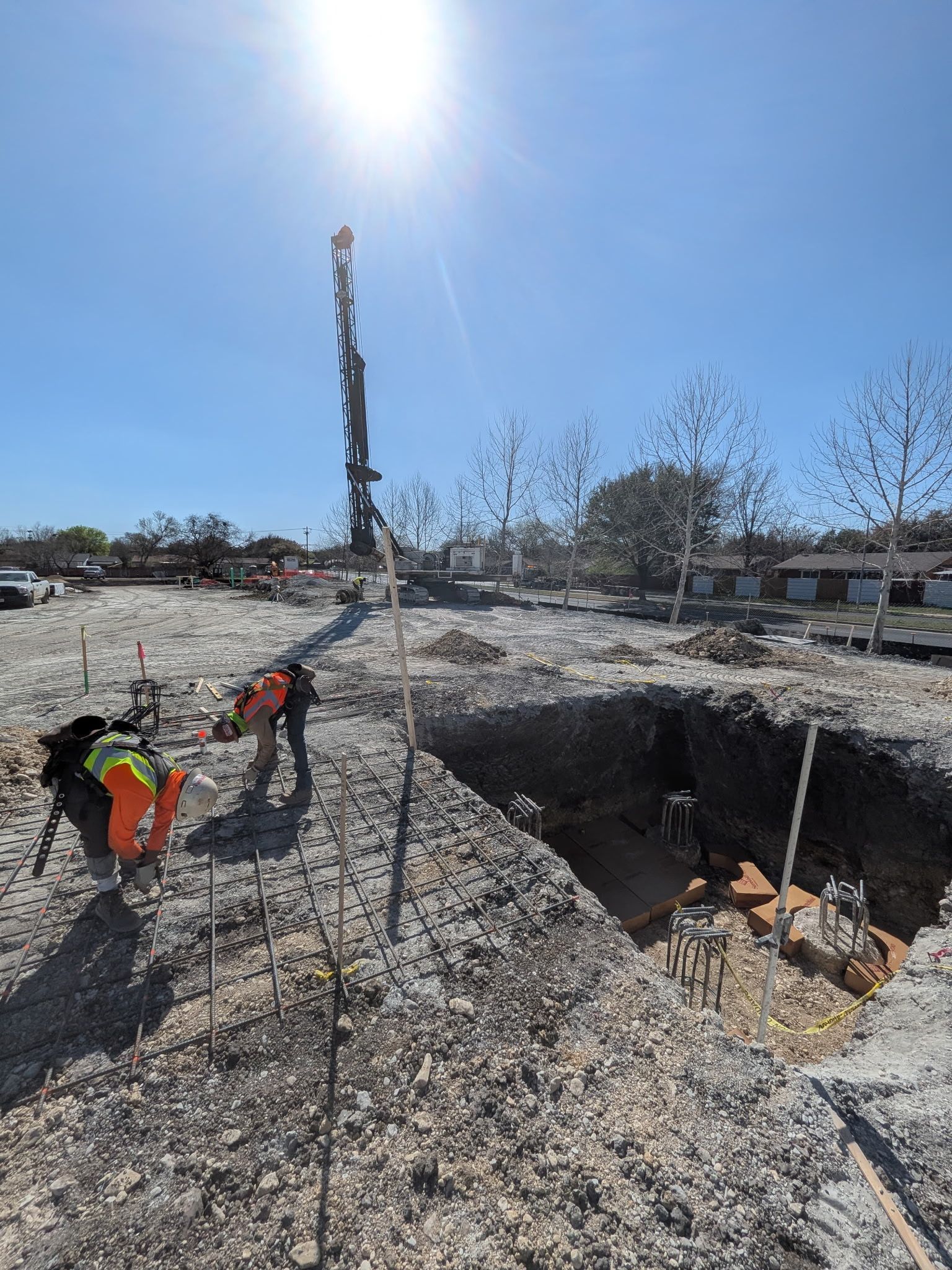 Construction workers in orange vests stand by a deep gravel pit under a bright sun.