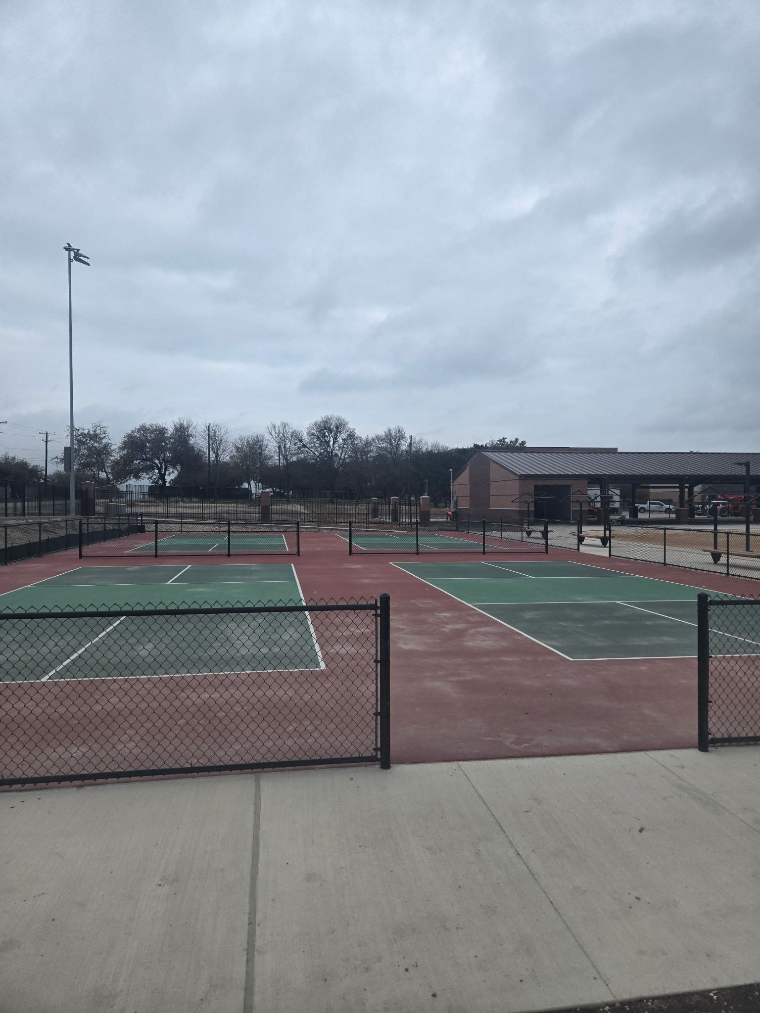 Outdoor tennis courts with green surfaces and red clay under a cloudy sky, seen from a fenced entrance.