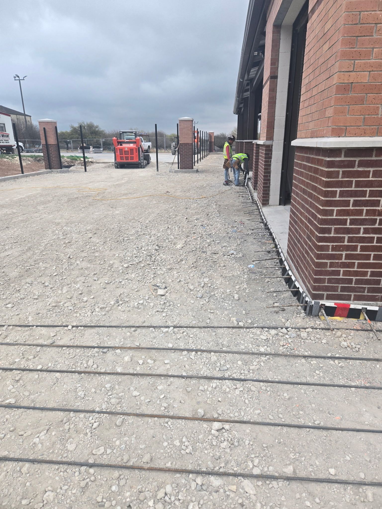 Gravel construction site beside brick building, with workers and a red machine in the distance.