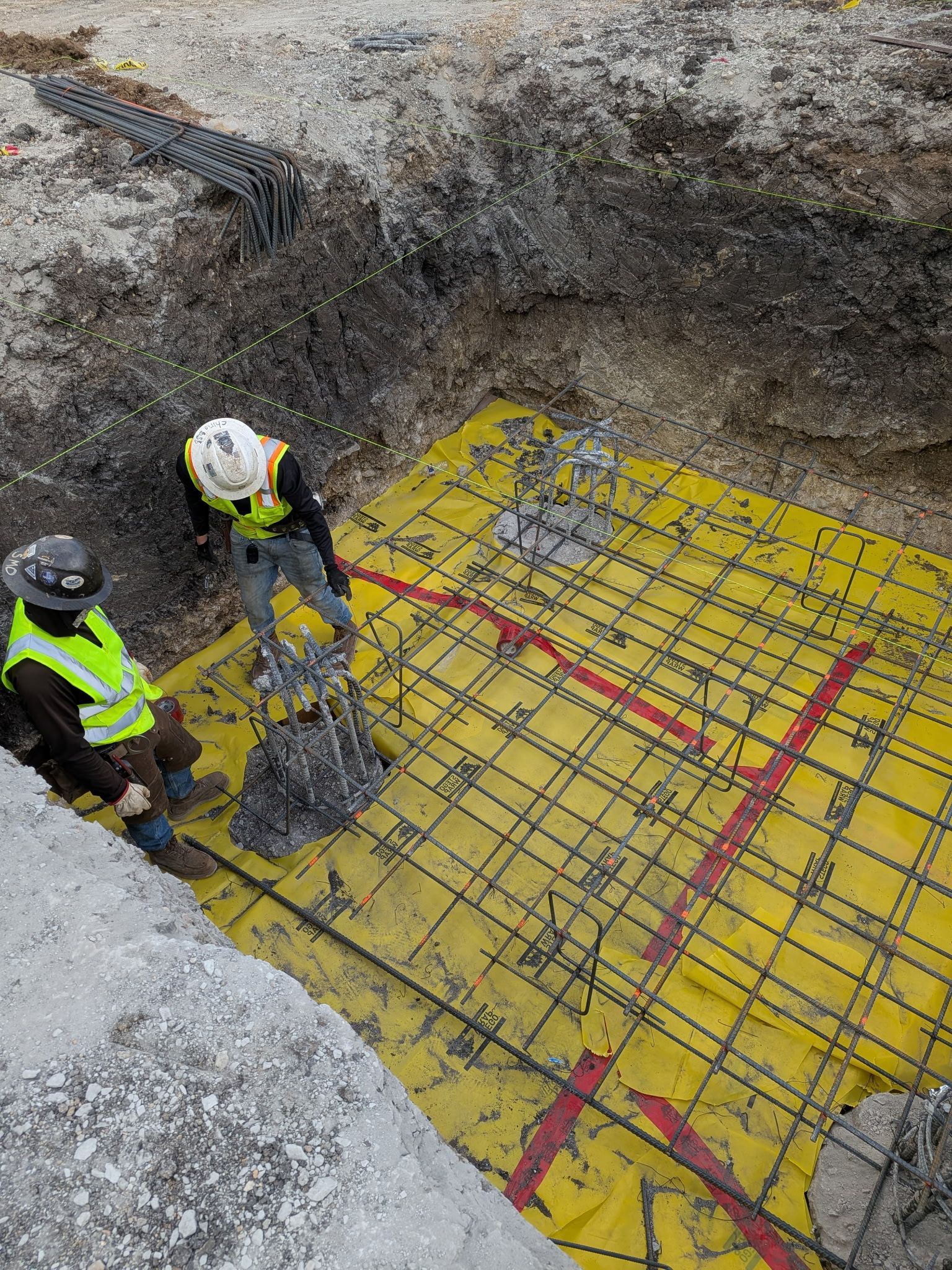 Two workers in safety vests inspect rebar over yellow waterproofing in a construction pit.