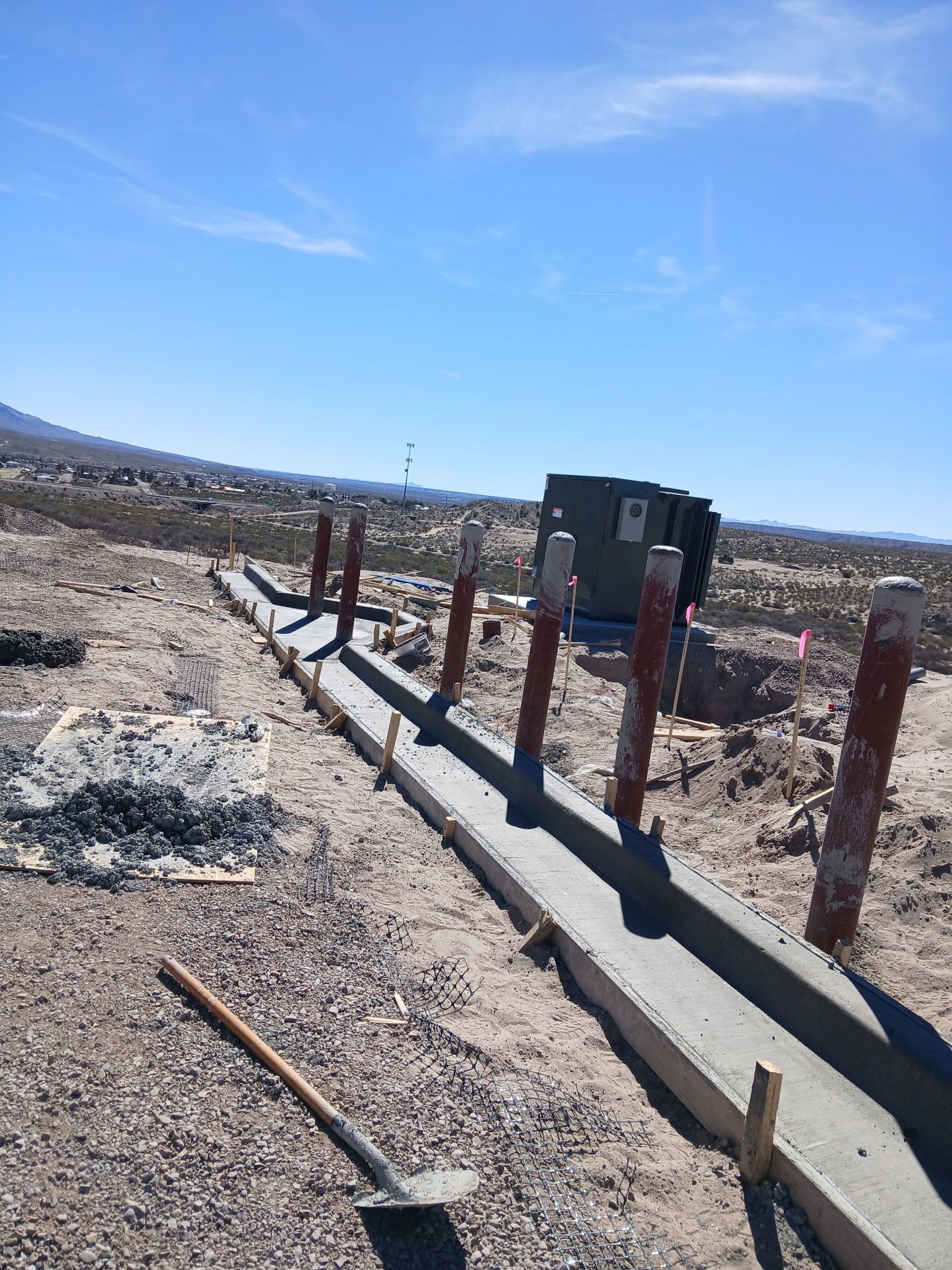 Rural roadside construction site with wooden posts, concrete curb, and utility boxes under a blue sky