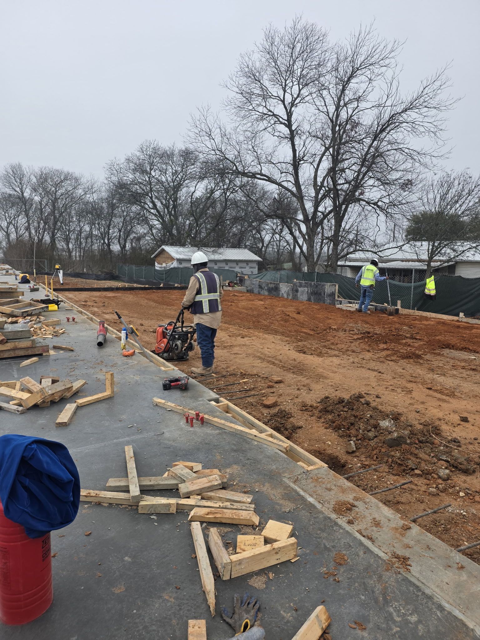 Roadwork scene with workers, orange dirt embankment, traffic cones, and scattered lumber beside a wet road