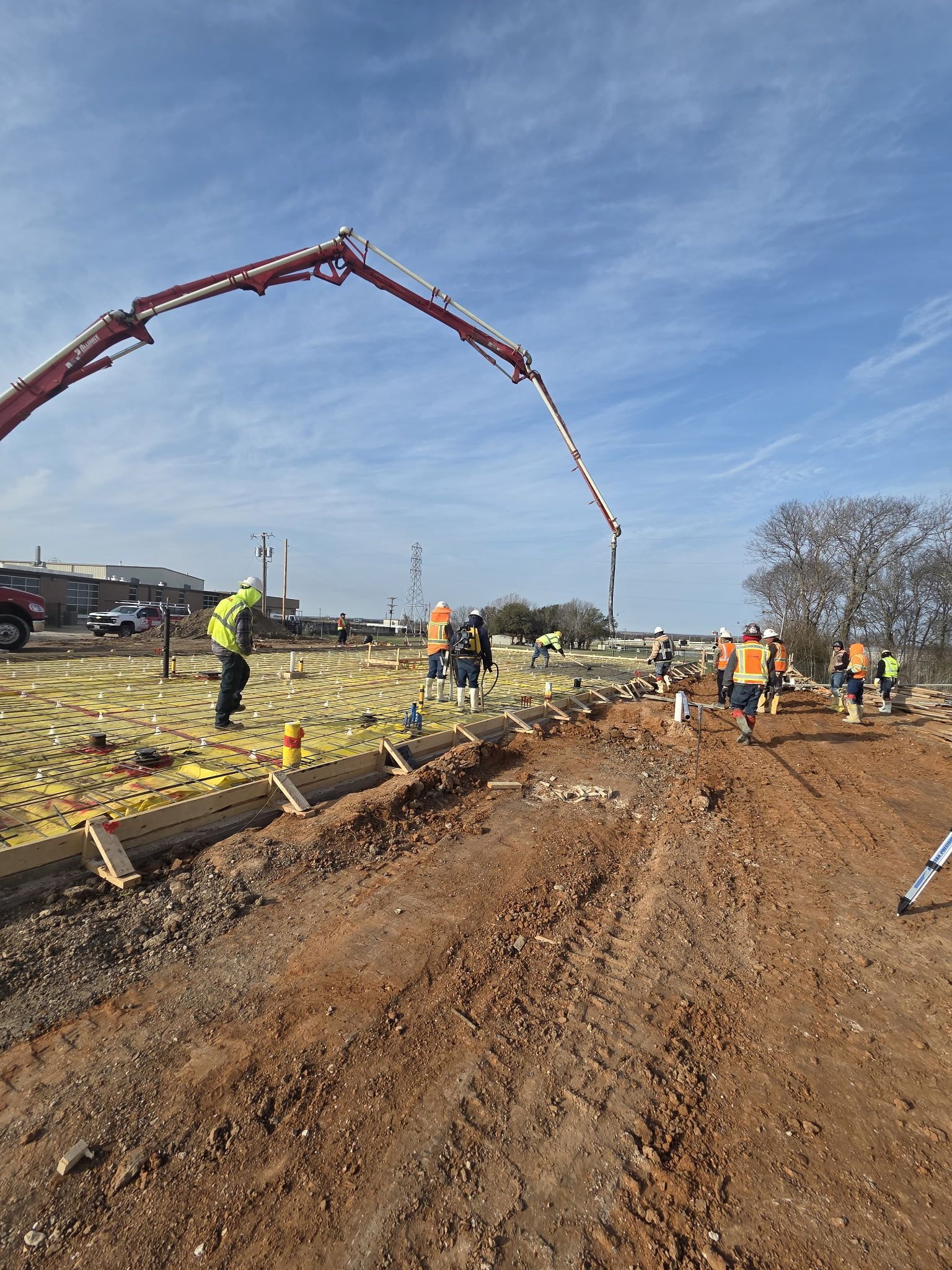 Construction crew pouring concrete on a road, with a long pump arm over a muddy site.