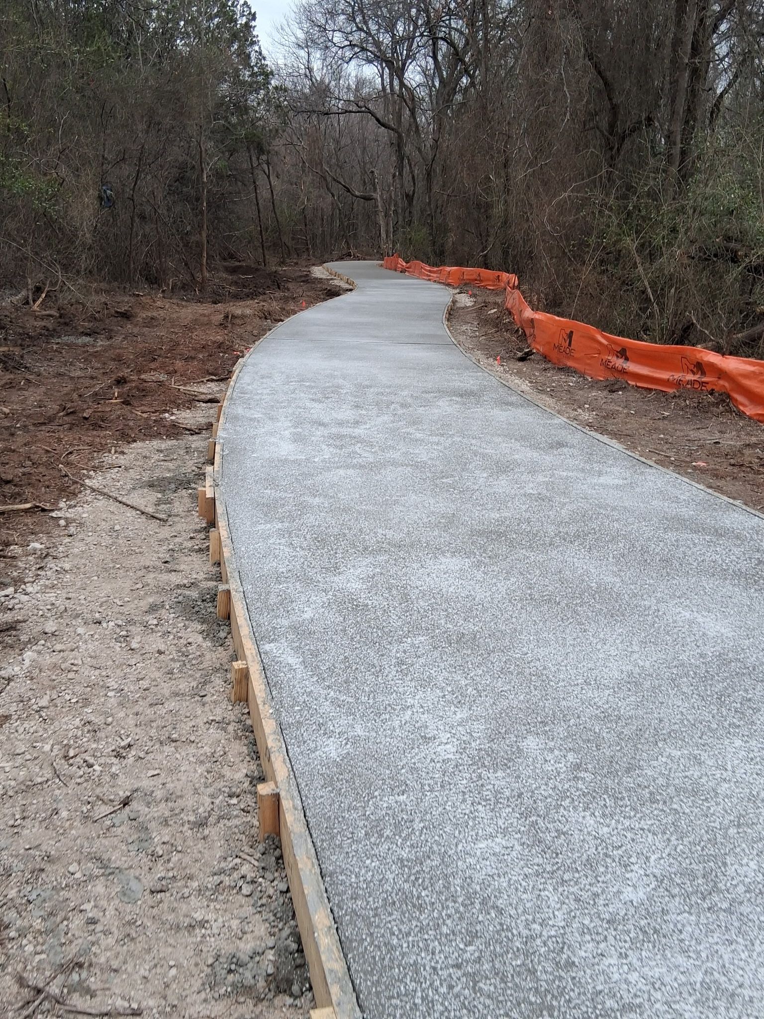 Curving gravel path under construction through woods, with orange safety fencing along the right side