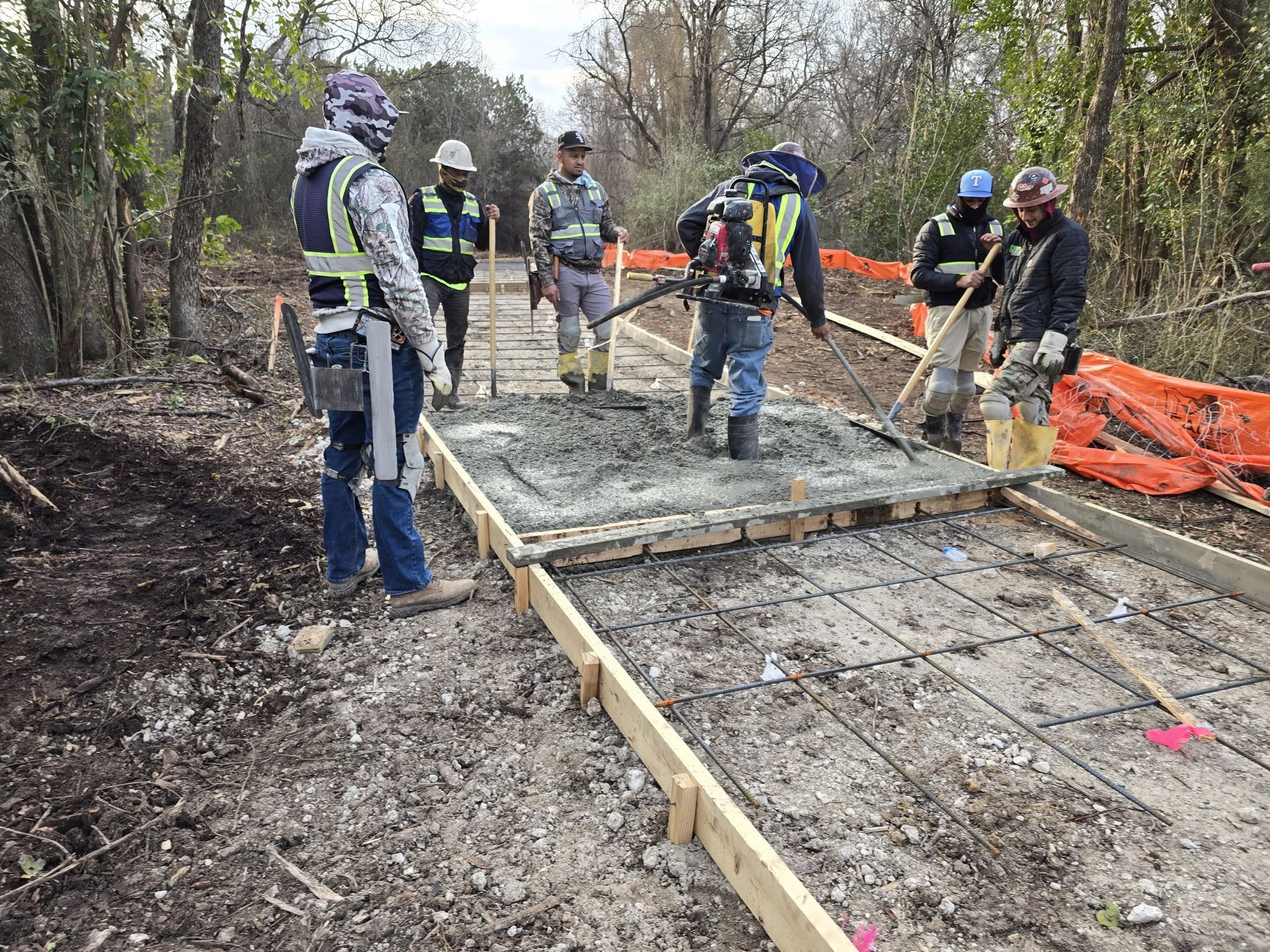 Construction workers pouring a concrete walkway in a wooded area, with forms and safety barriers.