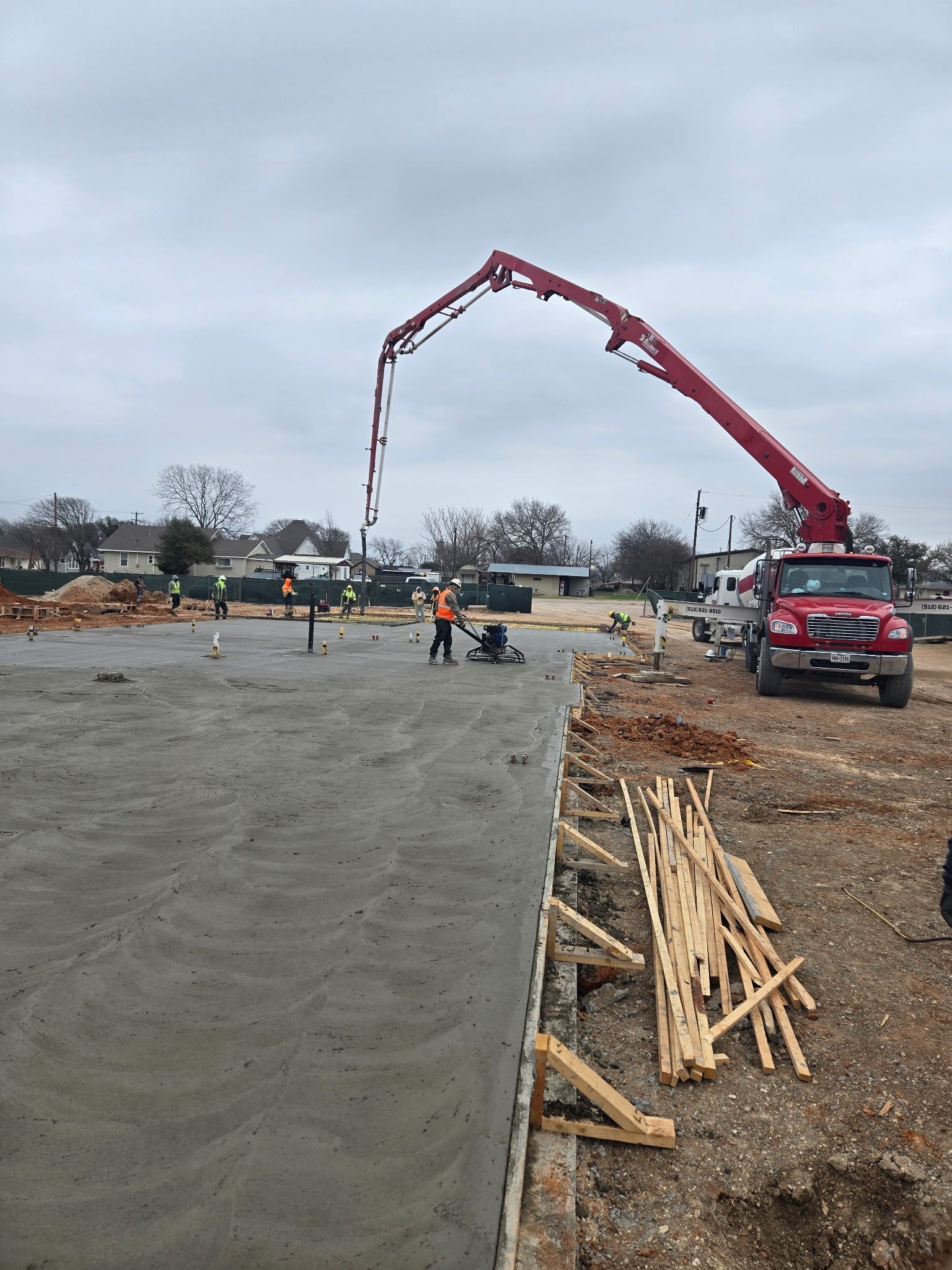Construction site with concrete truck pouring to a pavement area, workers nearby, and stacked lumber under a cloudy sky