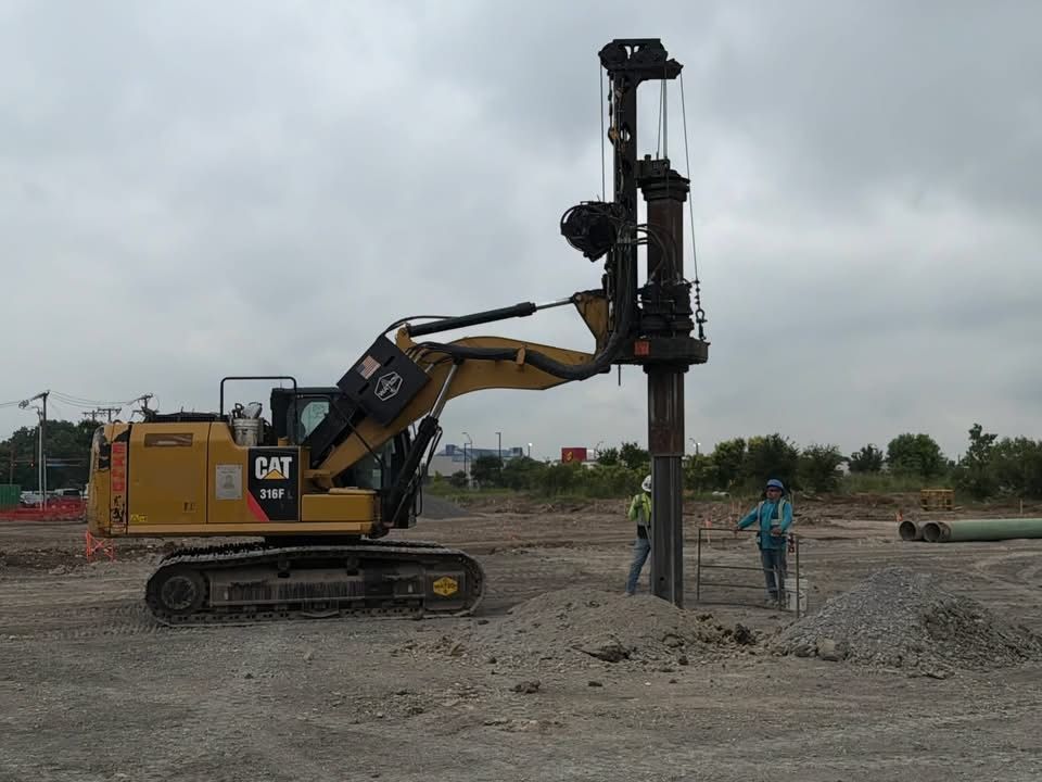 Yellow CAT excavator driving a drilling rig as workers stand beside it on a muddy construction site.