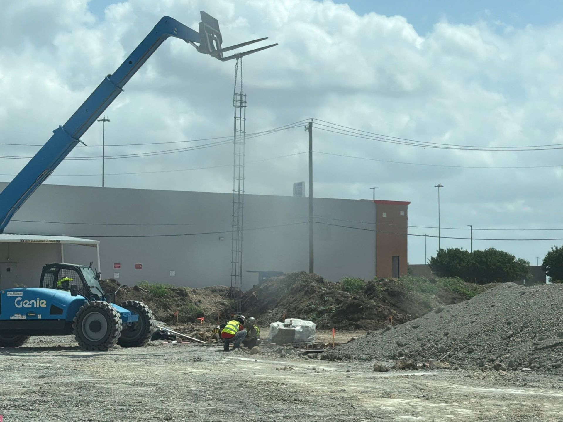 Construction site with a blue boom lift, worker, gravel pile, and large building under cloudy sky
