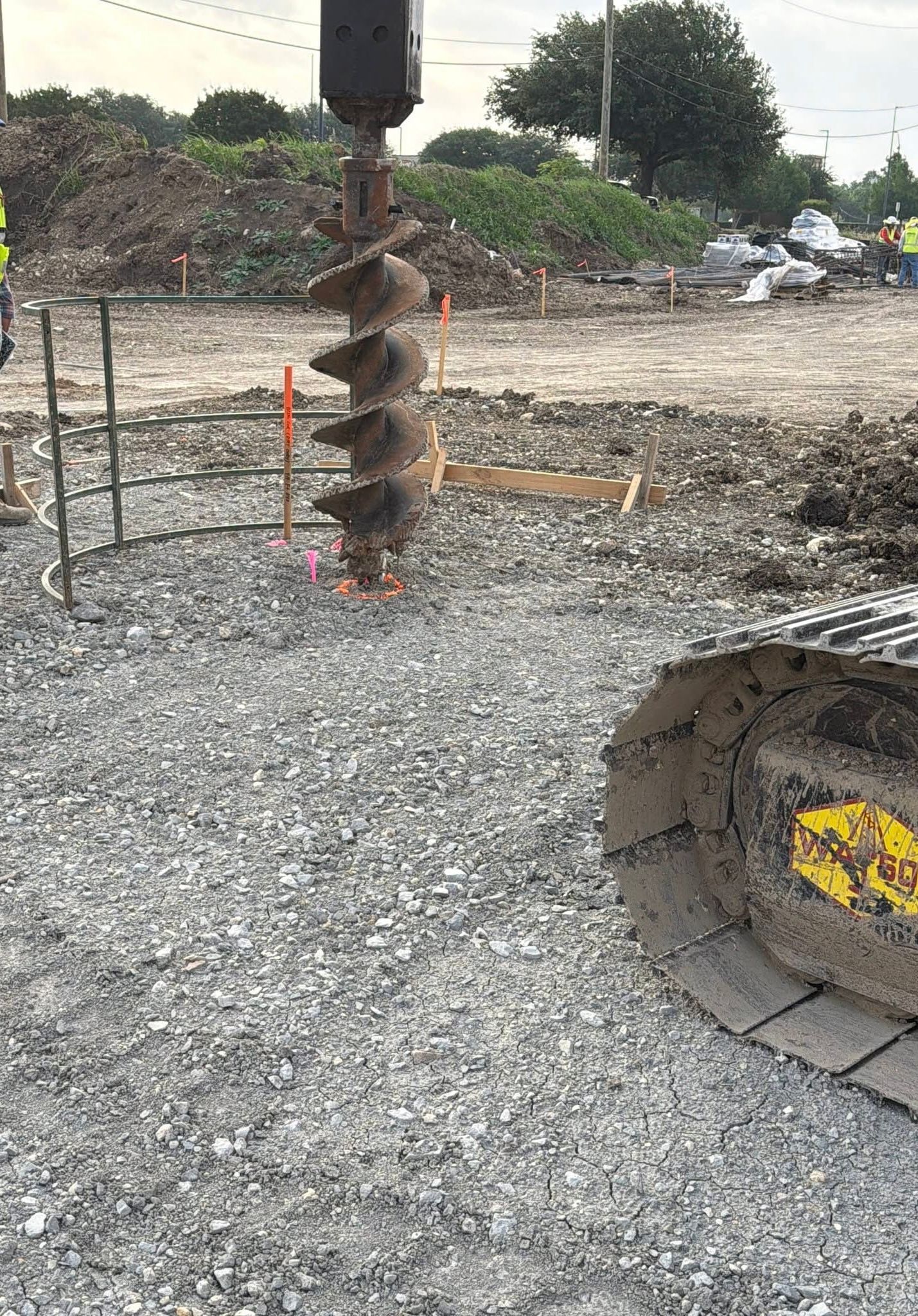 Large rusted drill bit over gravel at a construction site, with a concrete pipe opening nearby