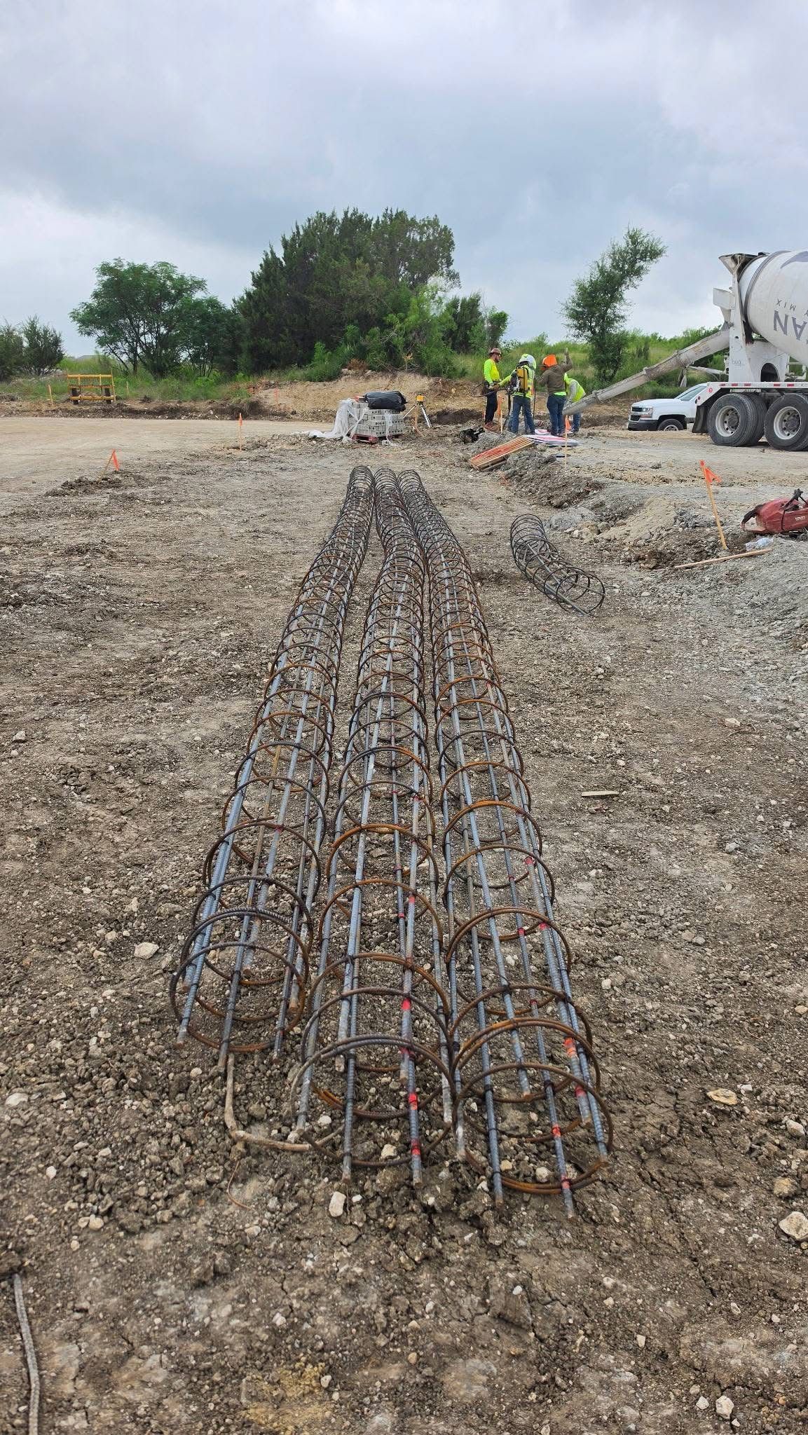 Rebar laid in a long trench on a gravel construction site with workers and a cement mixer in the background