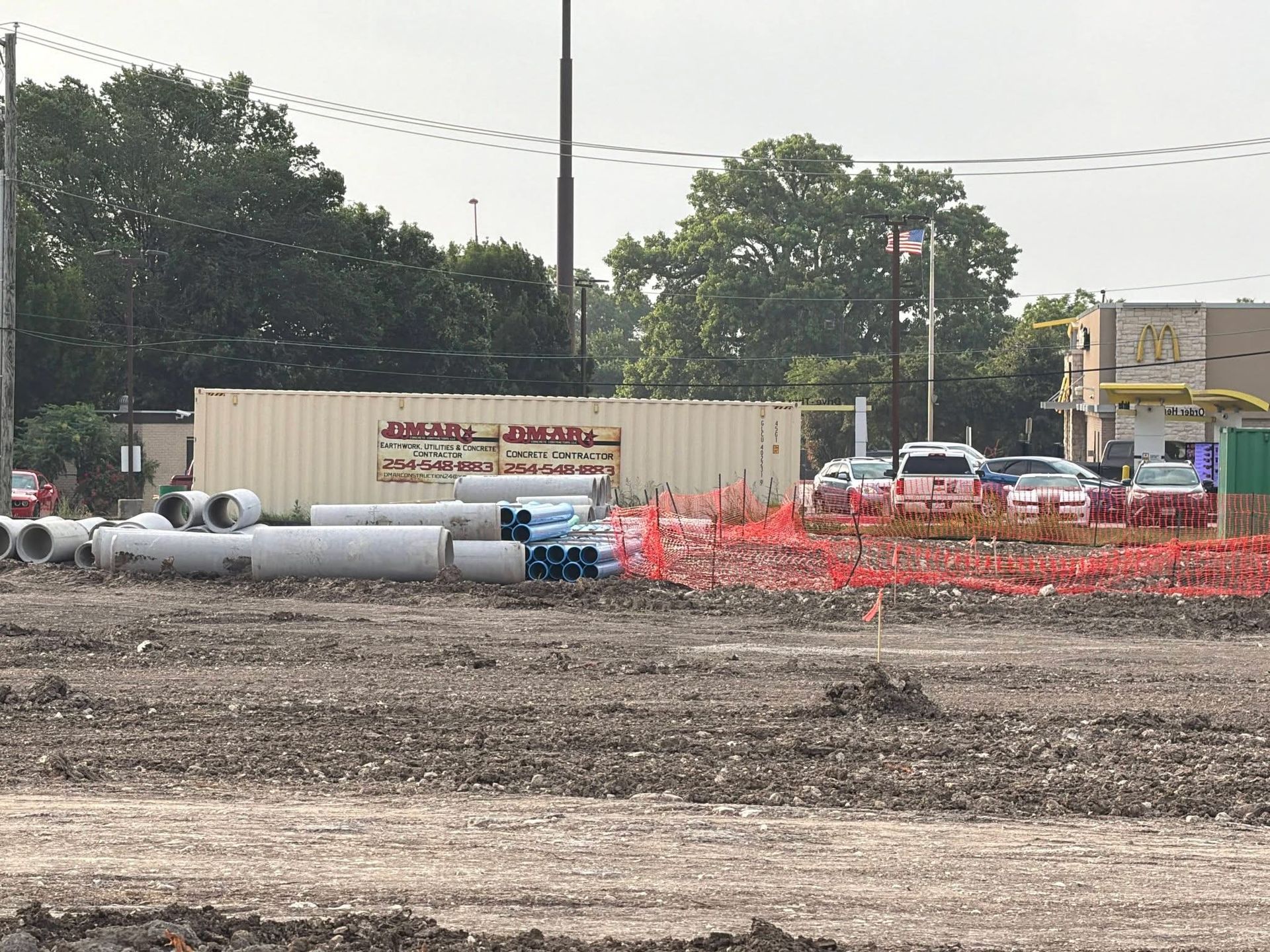 Construction site with pipes, orange safety fencing, shipping containers, and cars near trees and a road