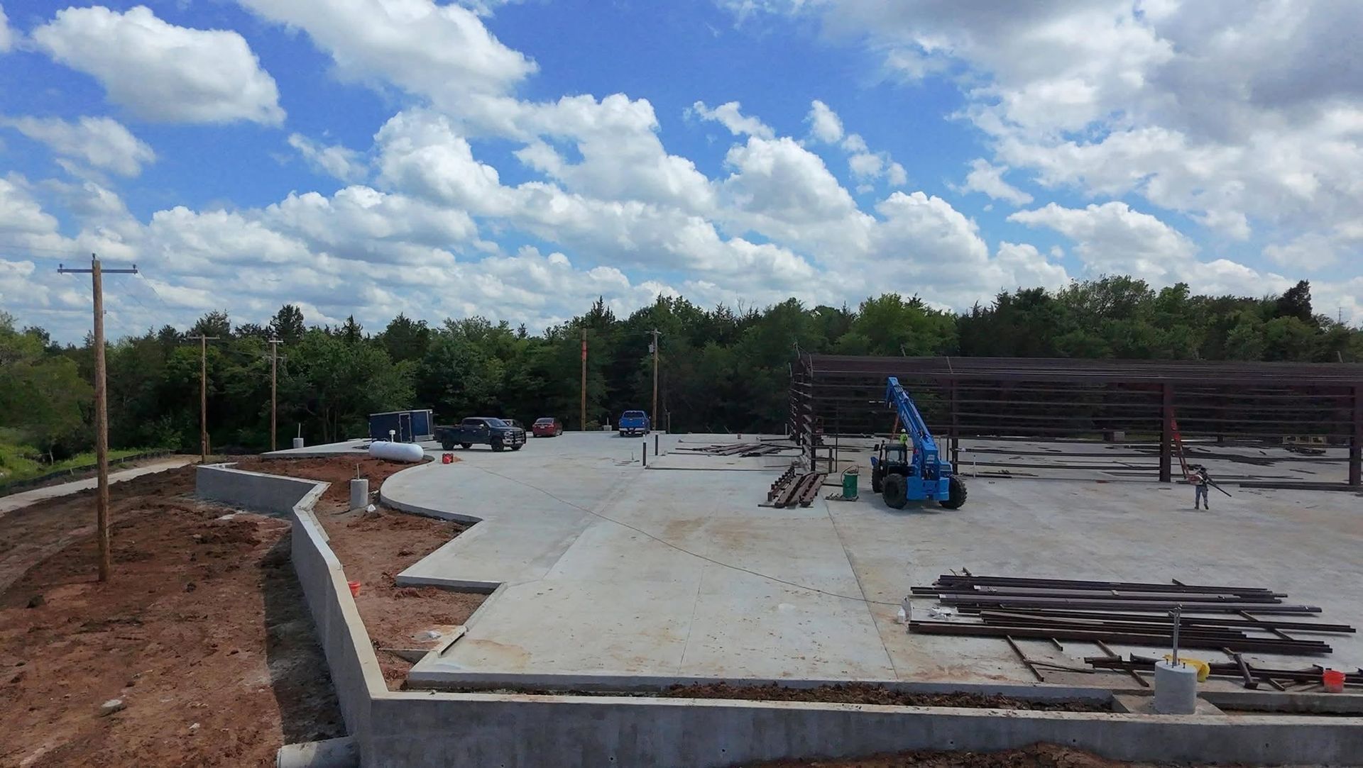 Concrete foundation under construction beside a wooded area, with steel beams and equipment nearby