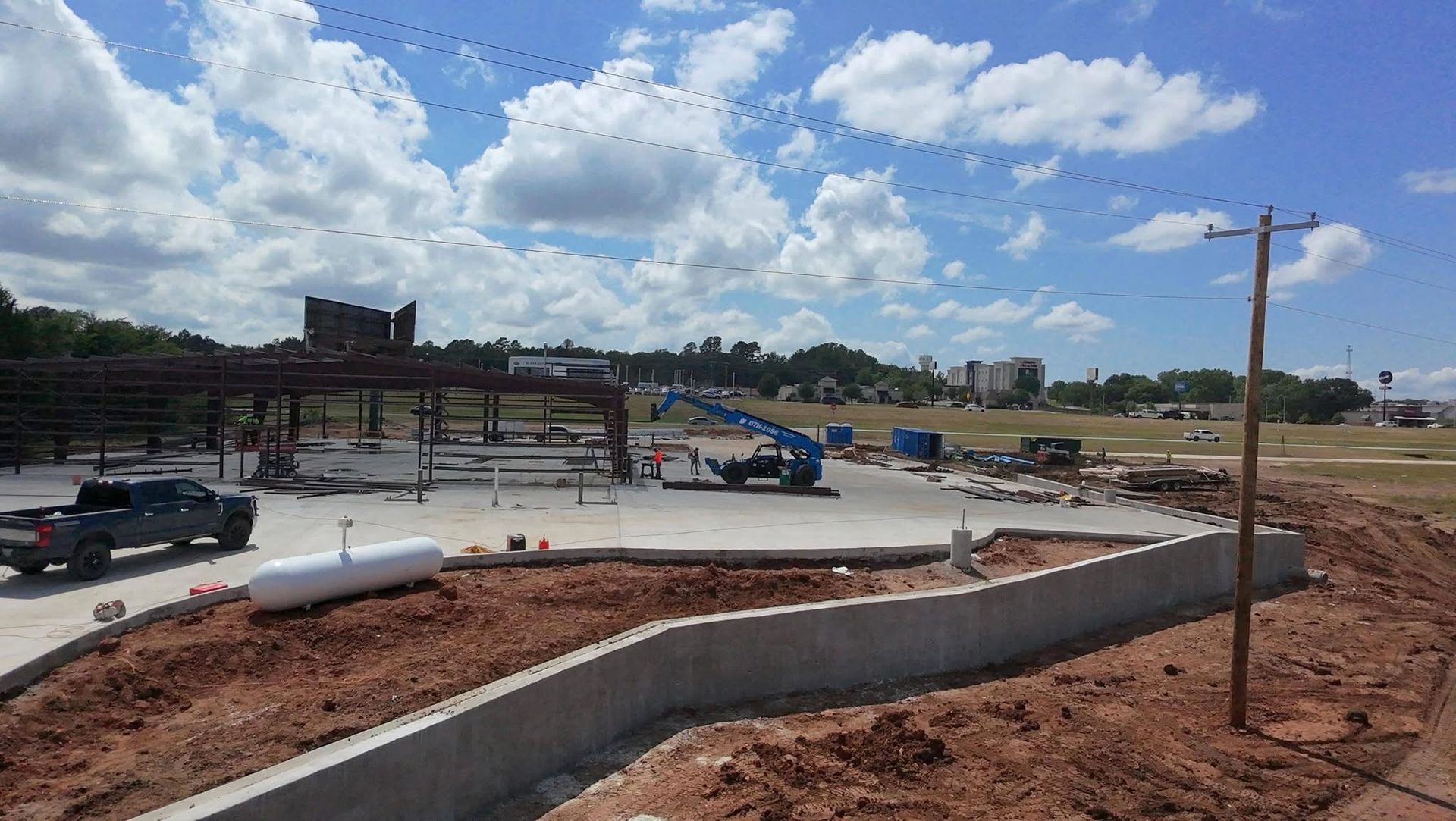 Construction site with concrete curb, red dirt, parked cars, and a partly cloudy sky