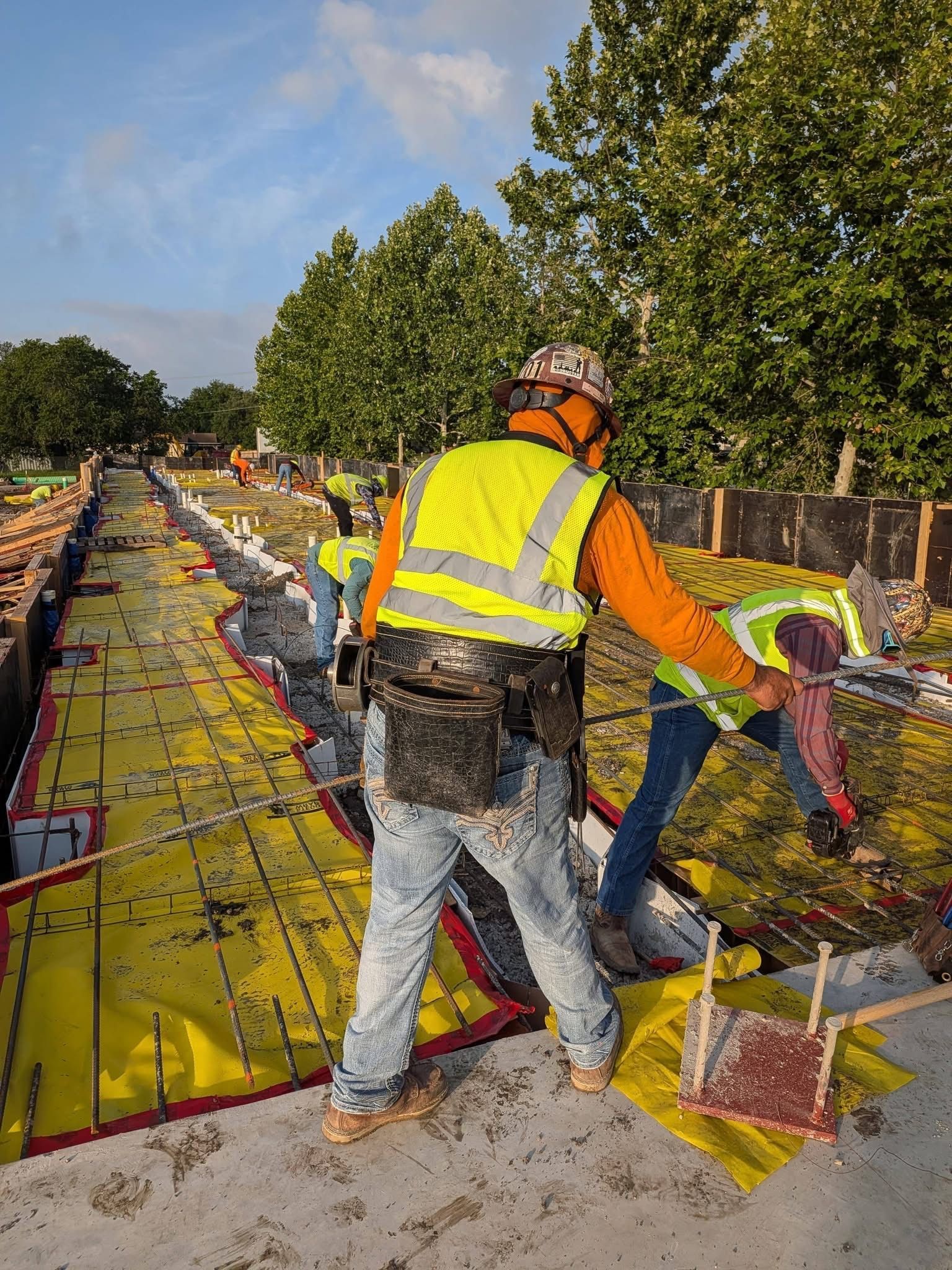 Construction workers in safety vests on a bridge deck, pouring concrete beside red formwork.