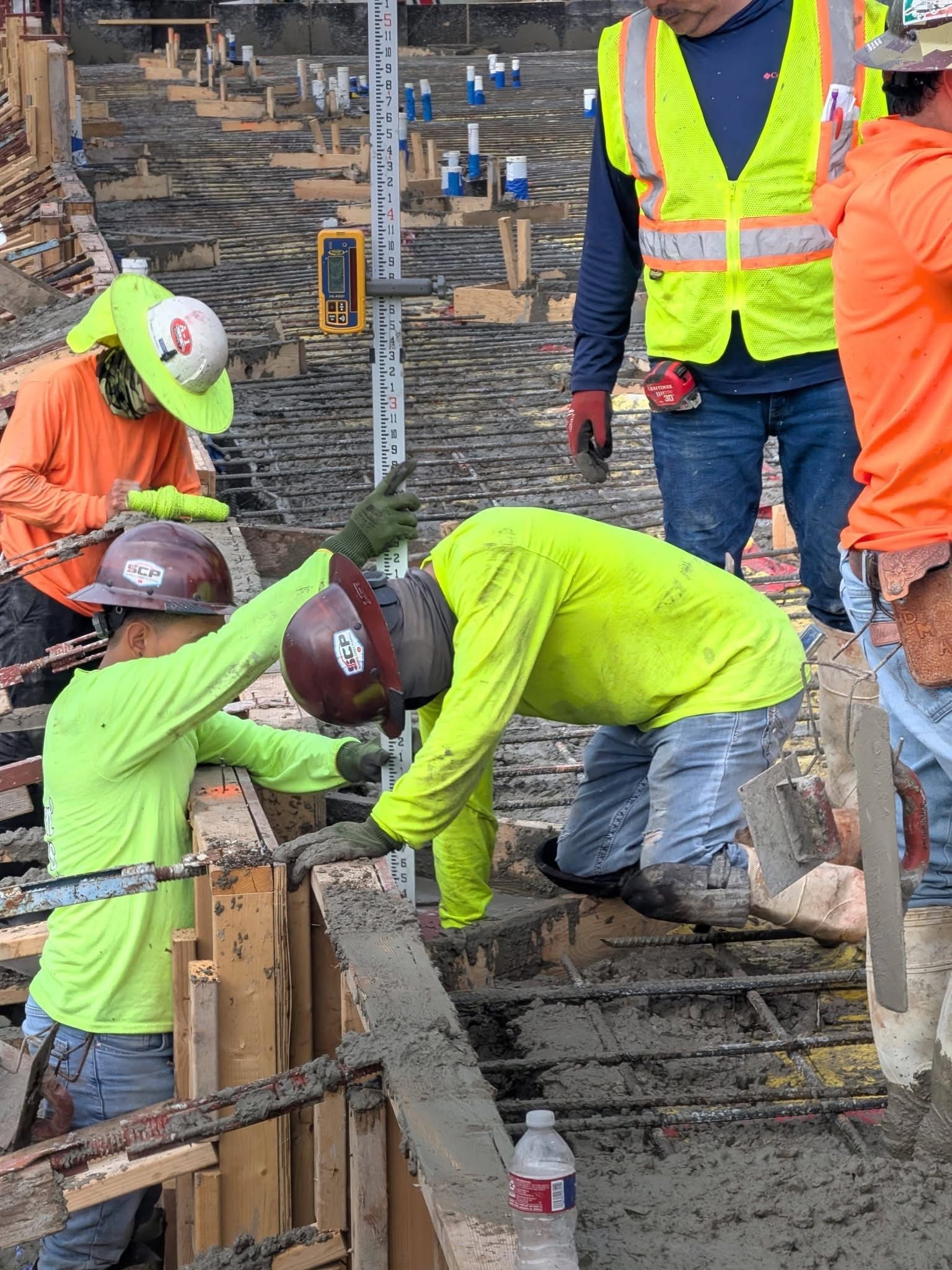Construction workers in neon safety gear working on concrete forms at a roadway site