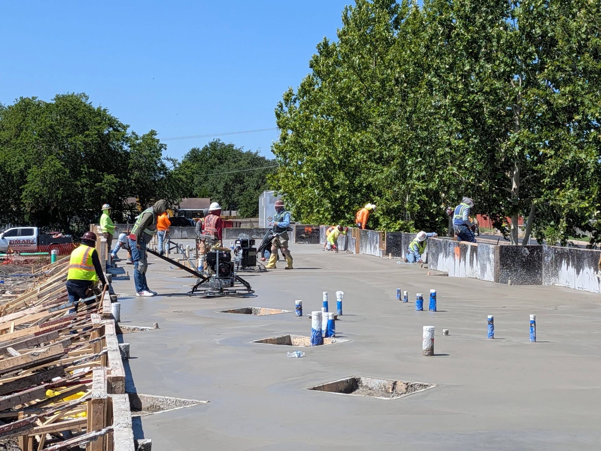 Workers paving a road section beside a line of concrete blocks and blue markers on a sunny day
