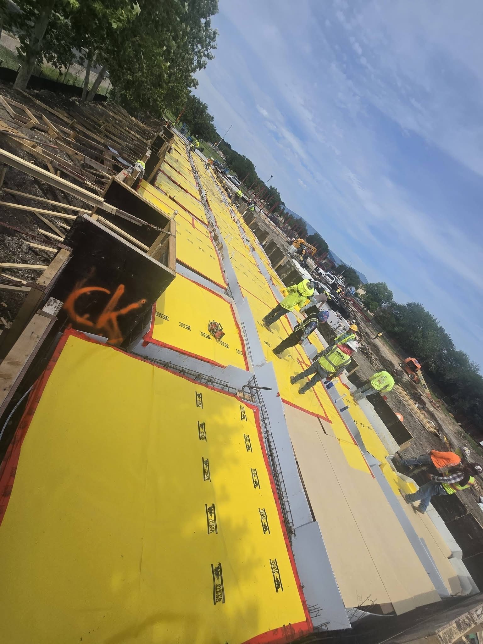 Workers installing yellow rooftop panels on a sloped roof beside a tree-lined street