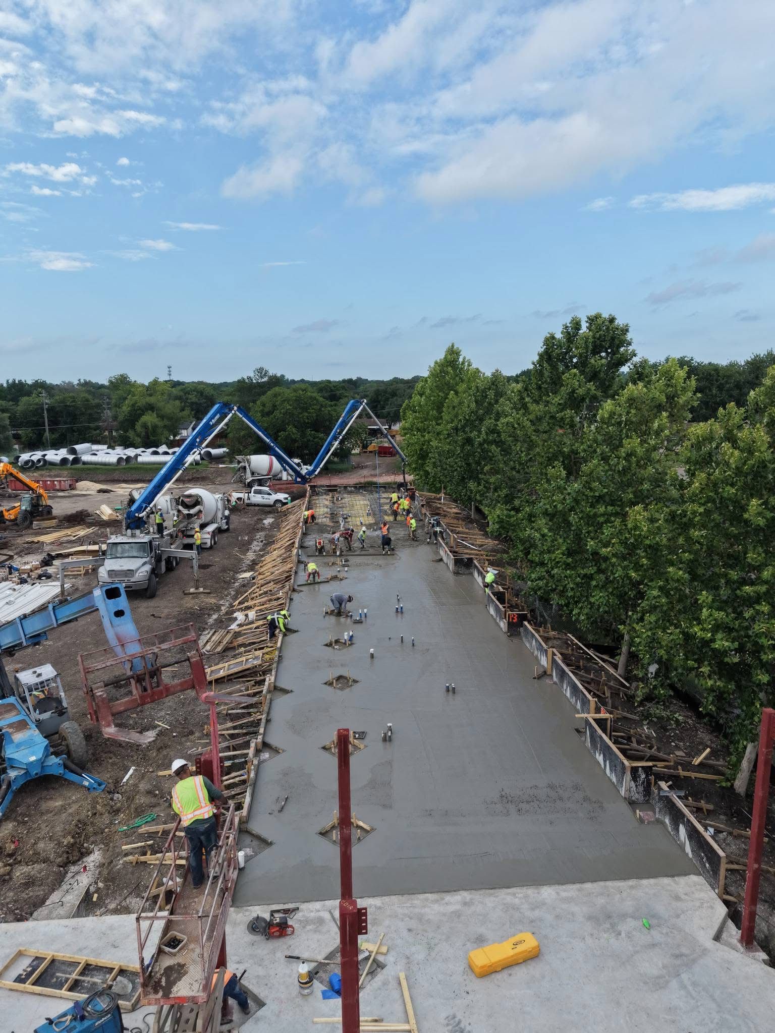 Construction workers excavating a long concrete channel beside trees and parked trucks under a blue sky