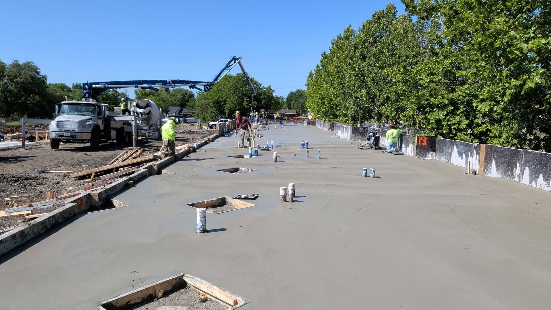Construction crew pouring concrete on a wide road beside trees and parked vehicles
