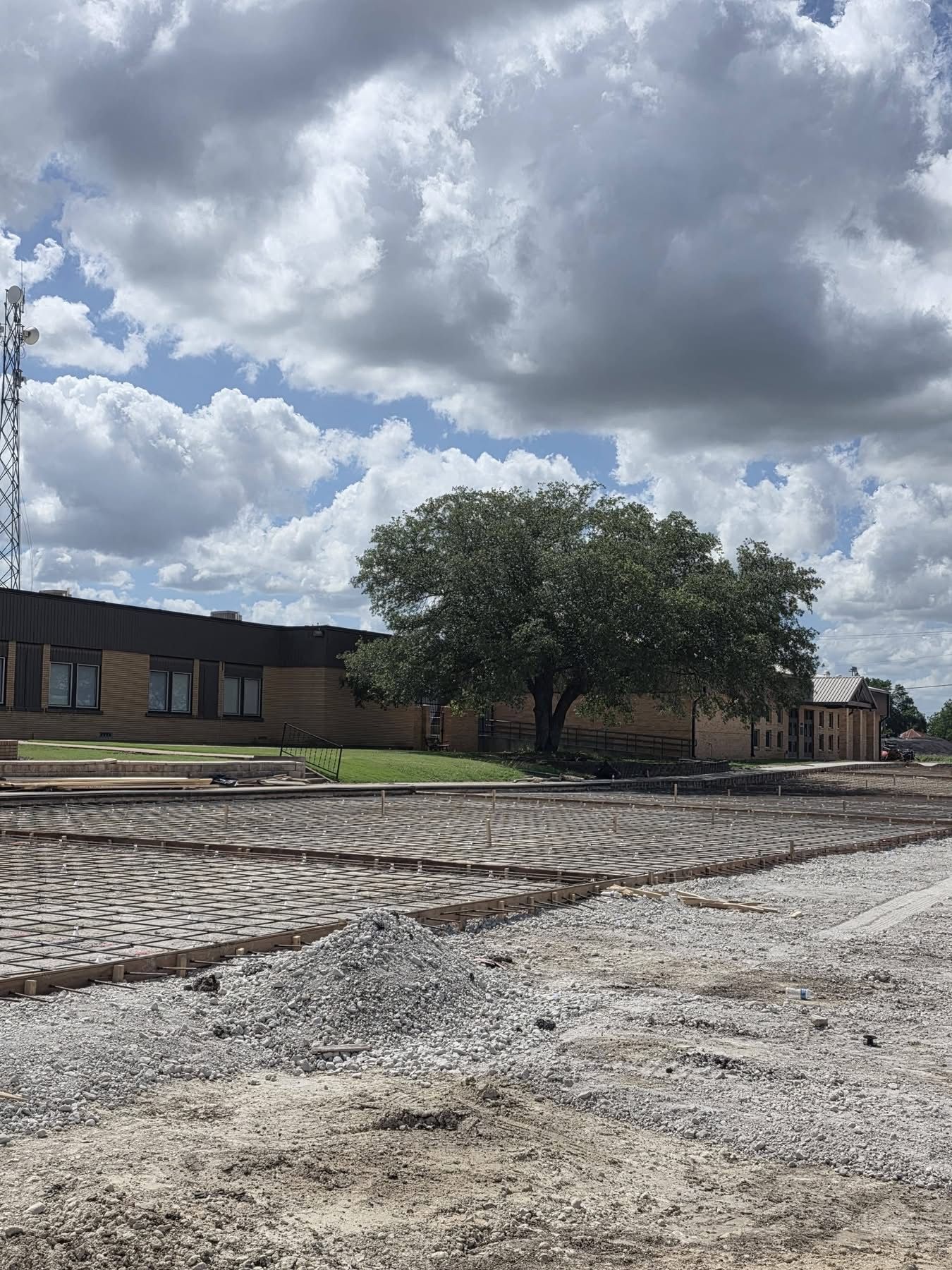 Gravel lot under cloudy sky beside low brick buildings and a large tree