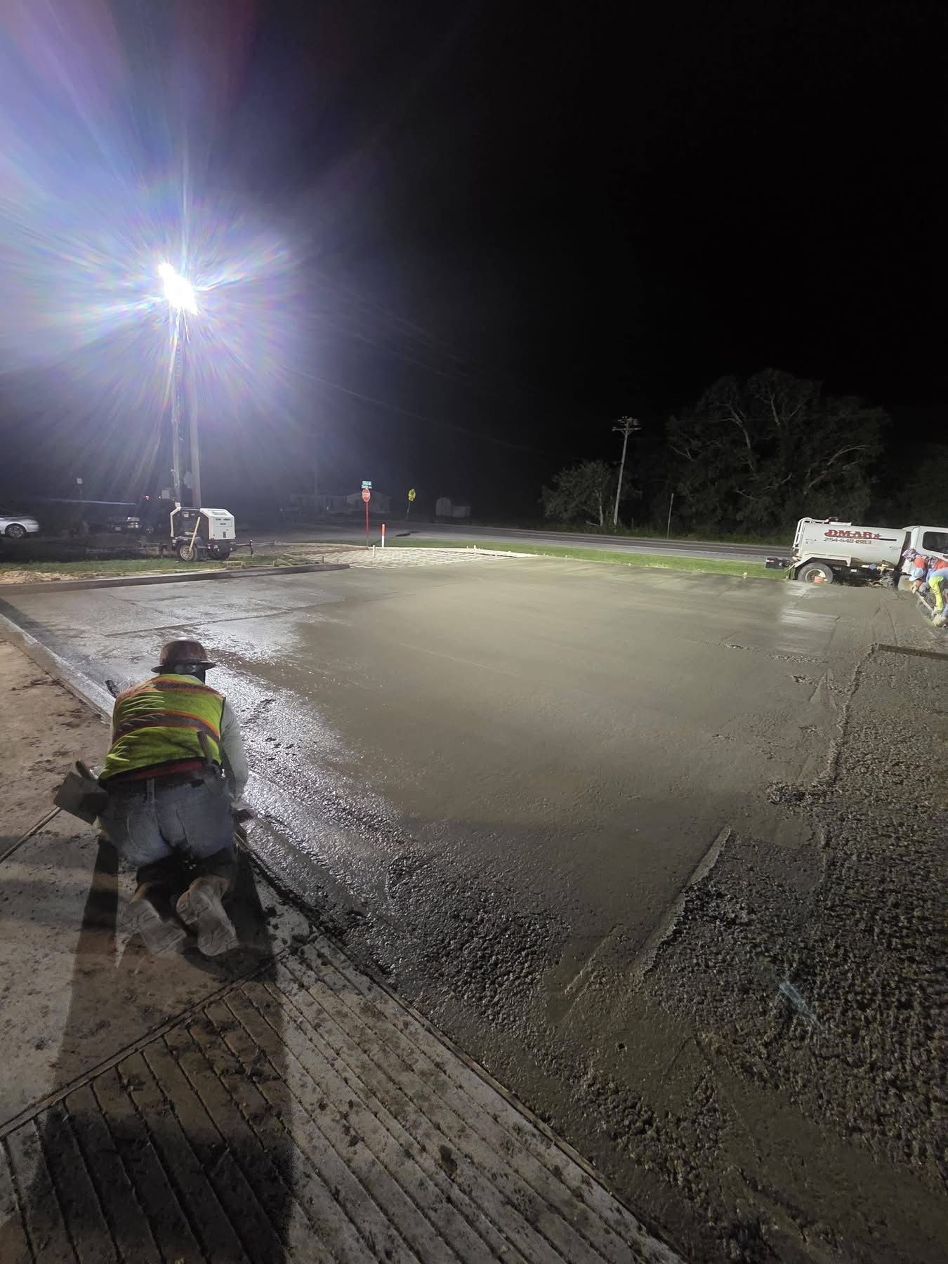 Worker crouching on a wet road at night beside a bright streetlight and construction equipment