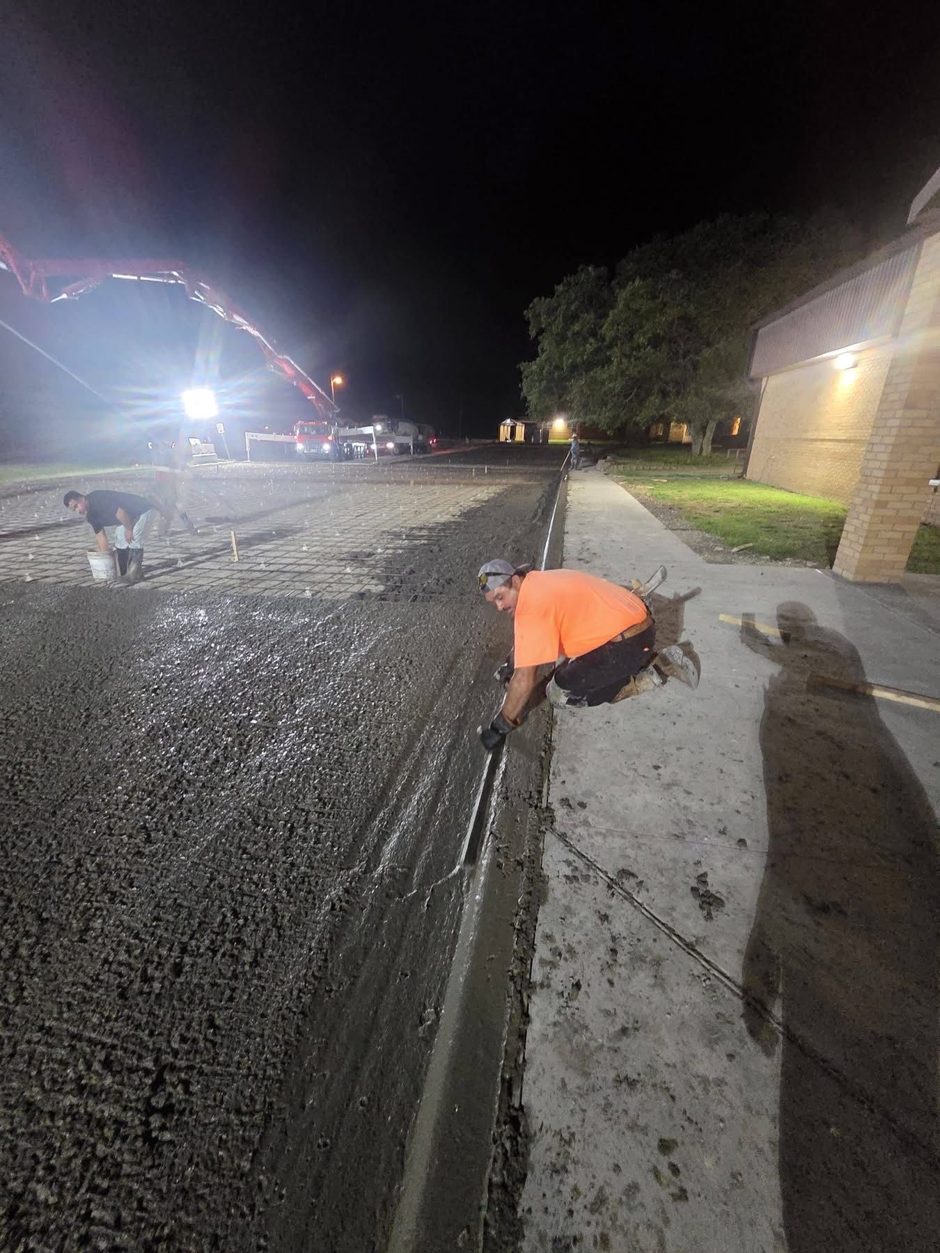 Workers paving a road at night, one in an orange jacket kneeling by the curb.