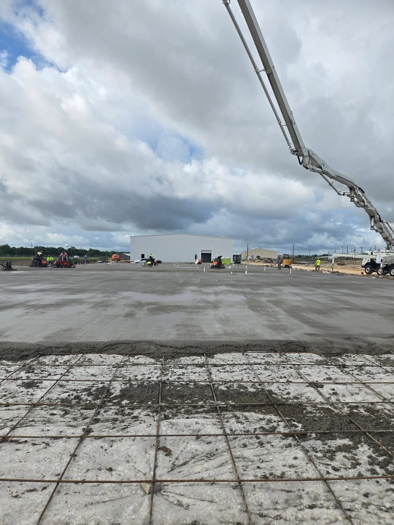 Concrete worksite under cloudy sky with a pump boom extending from the right and wet ground in the foreground