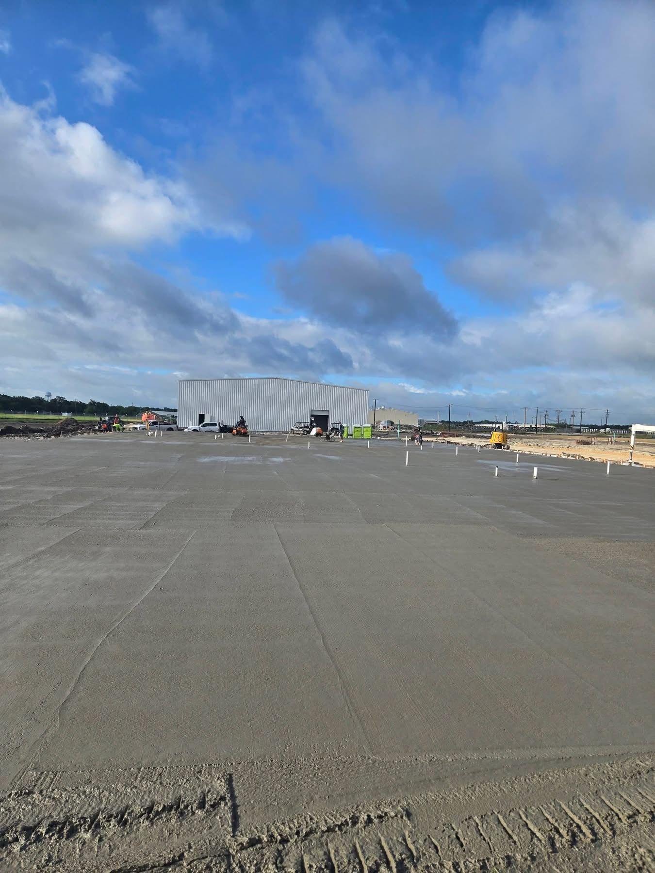 Wide empty tarmac with a large white building under a partly cloudy blue sky, people and equipment in the distance