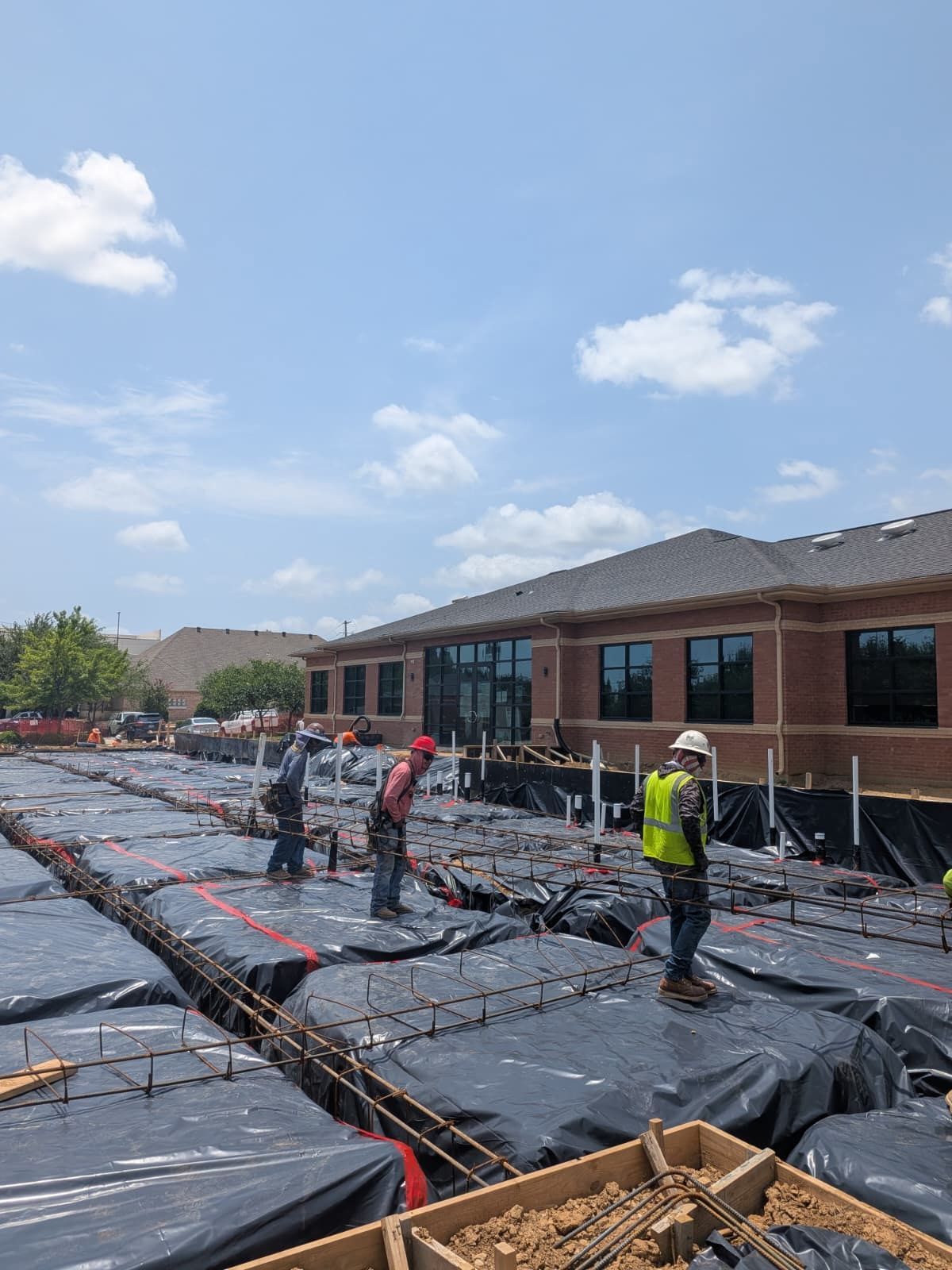 Workers installing rebar and black waterproofing on a large construction slab beside a brick building
