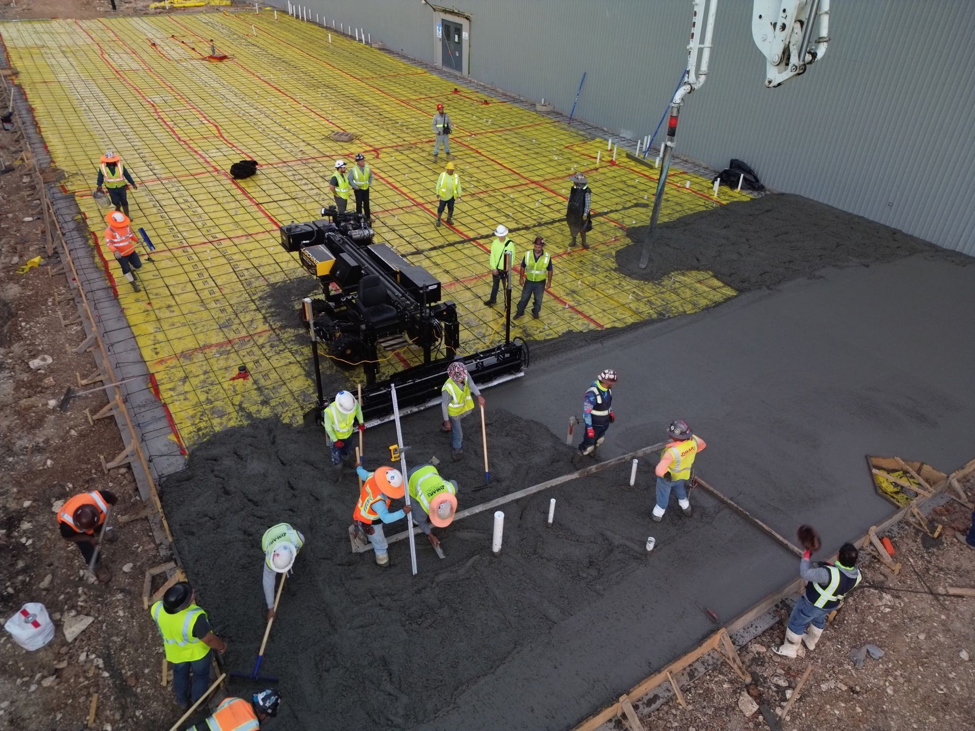 Construction crew paving a large asphalt area around heavy equipment and yellow rebar mats in a worksite.