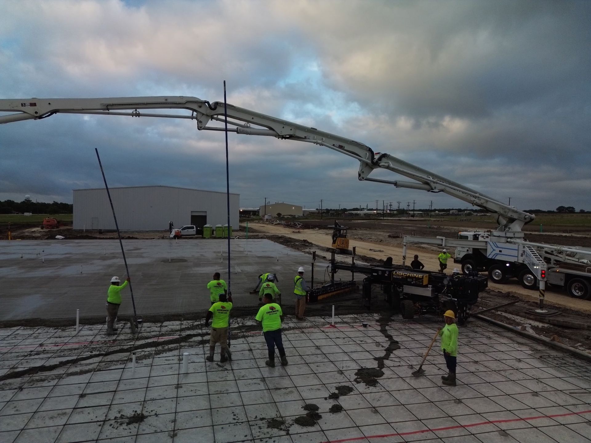 Workers in neon vests operate a concrete pump at a construction site under cloudy skies.