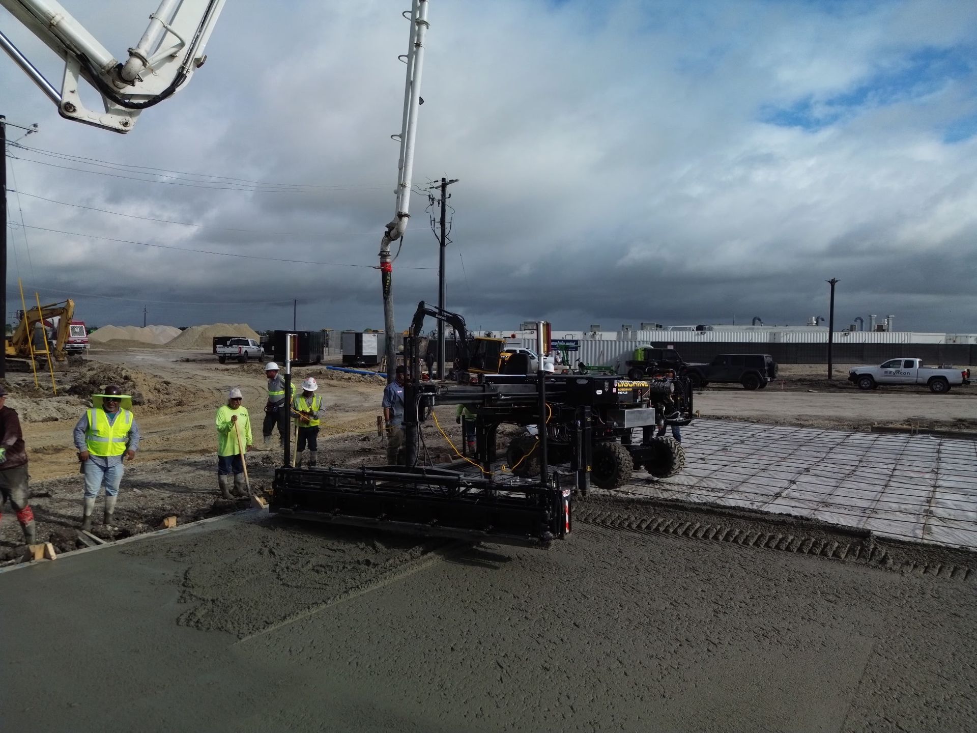 Road paving crew working on a wet roadway under cloudy skies, with a paver and workers in high-visibility vests.