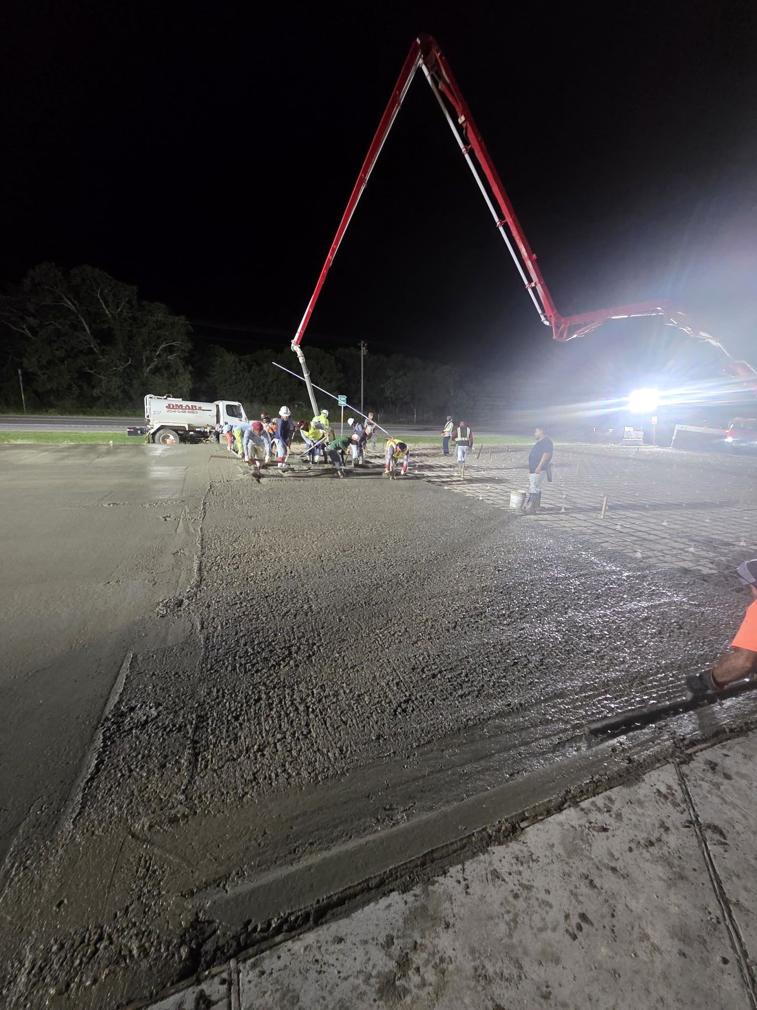Night construction site with a crane lifting a red-lit structure over gravel and workers nearby.