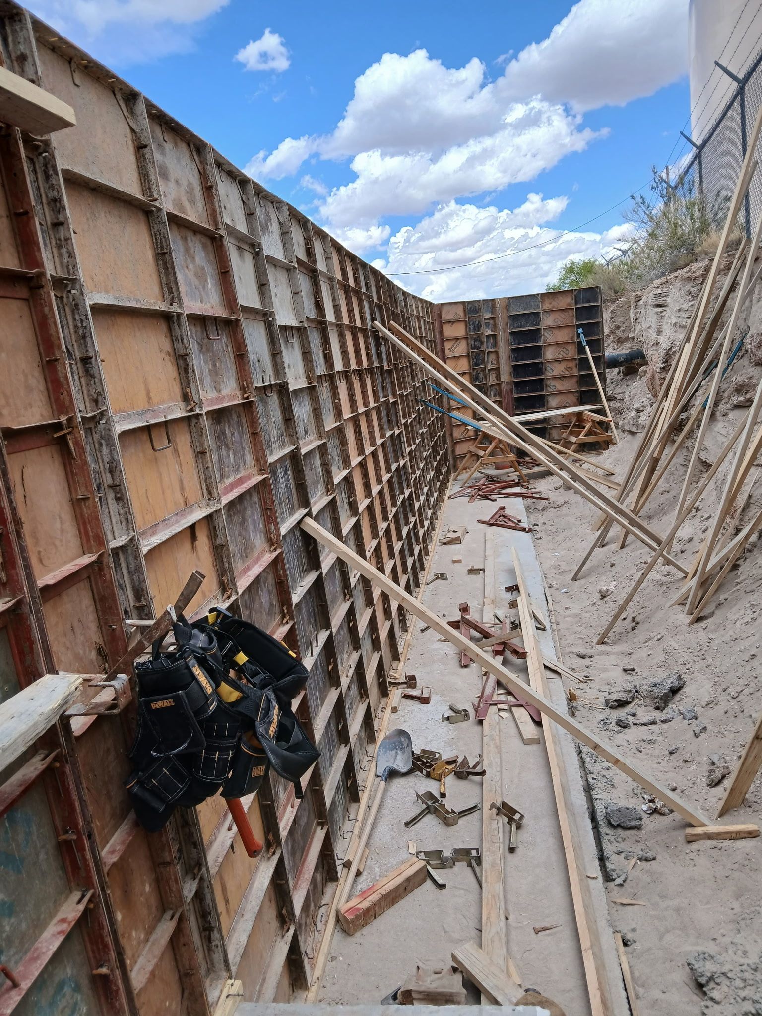 Construction site trench with wooden formwork, metal braces, and scattered debris under a blue sky.