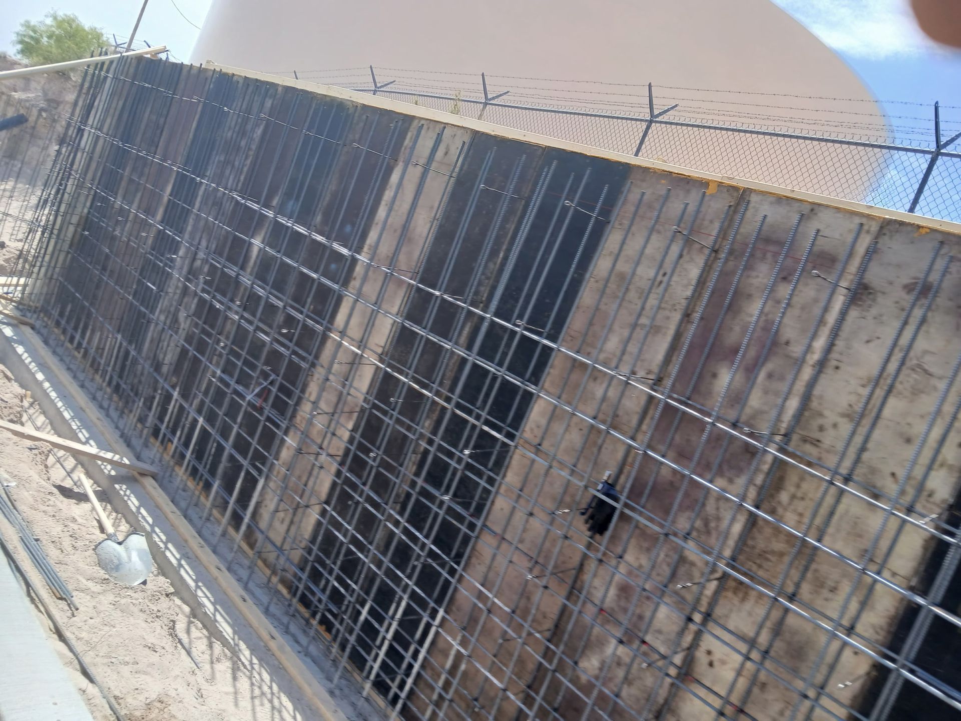 Tilted wire-mesh panels leaning against a concrete wall with barbed wire on top in a dusty outdoor area