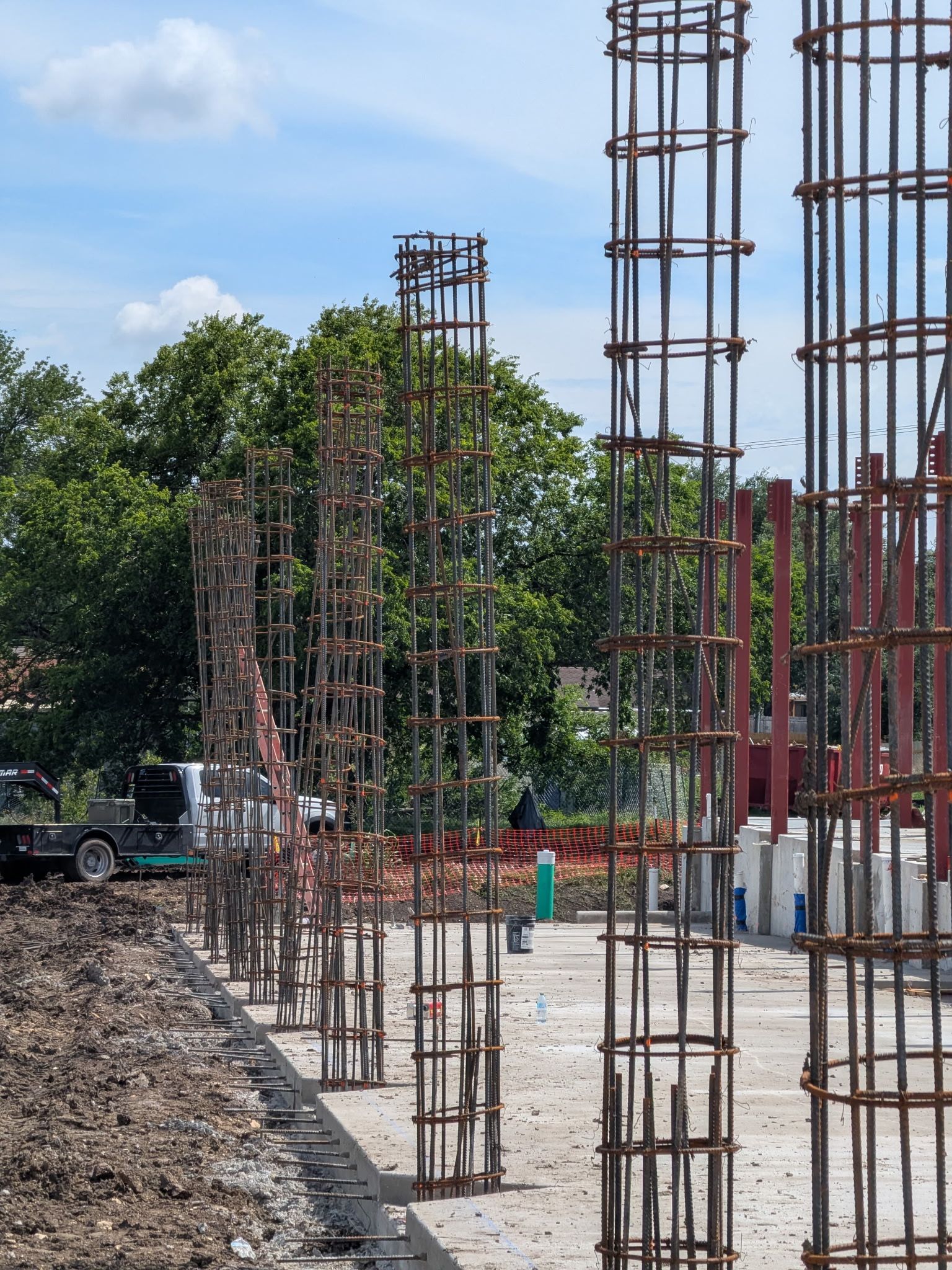 Vertical steel rebar columns at a construction site beside a gravel road and trees under a blue sky