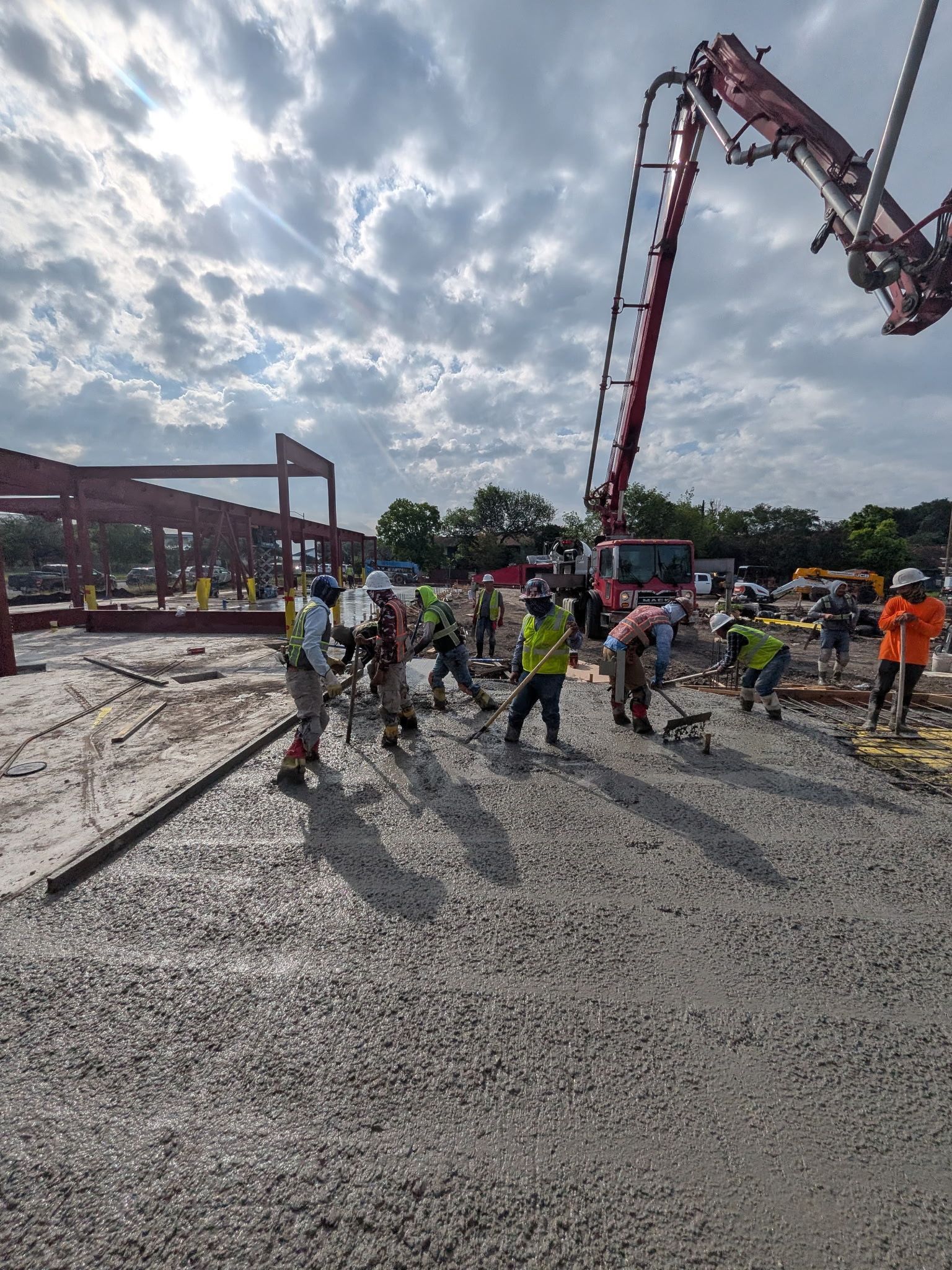 Construction crew pours concrete at a building site under a cloudy sky, with a boom pump overhead.