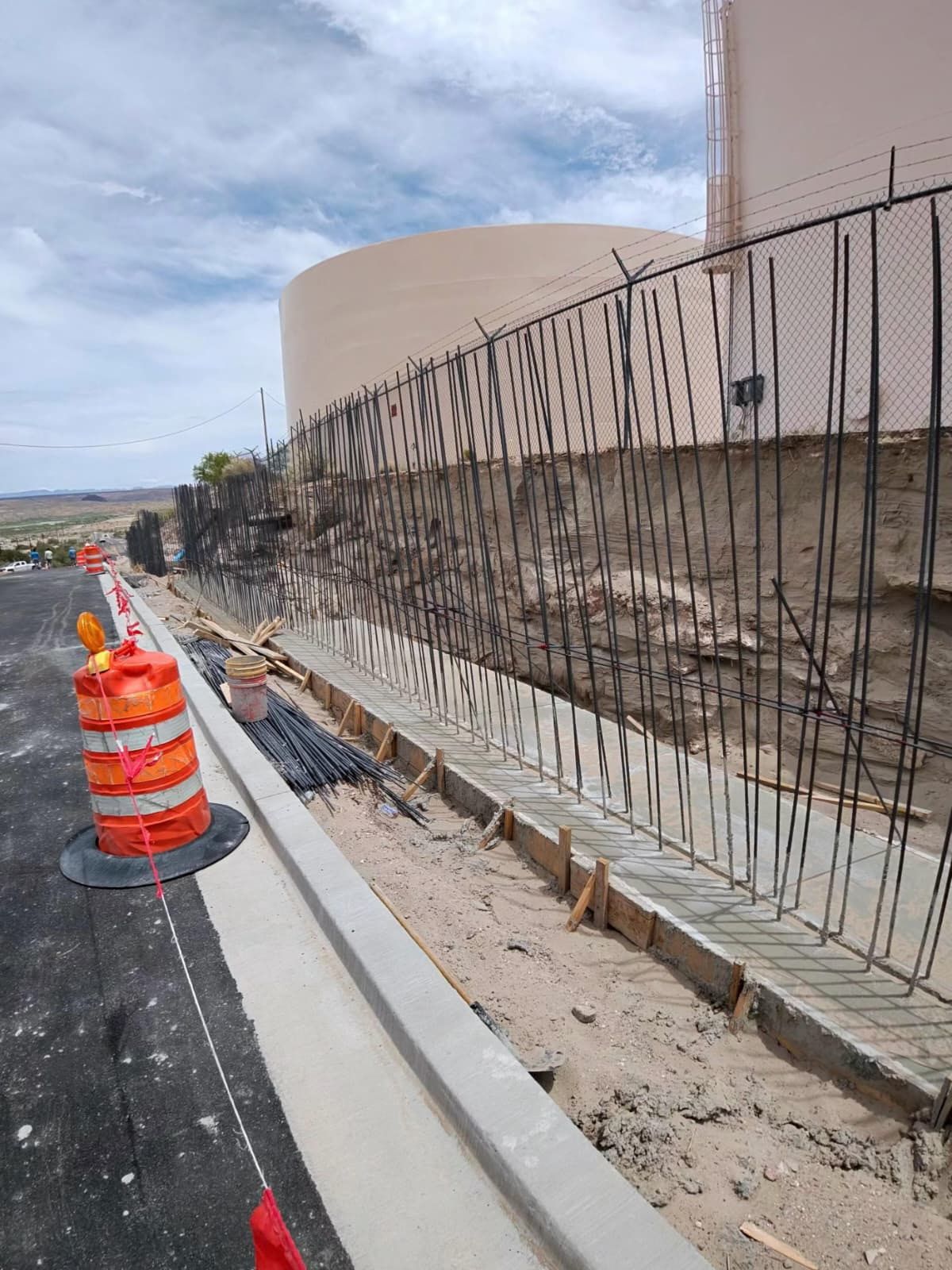 Construction site with orange traffic barrel, curb, and exposed rebar beside a large concrete structure