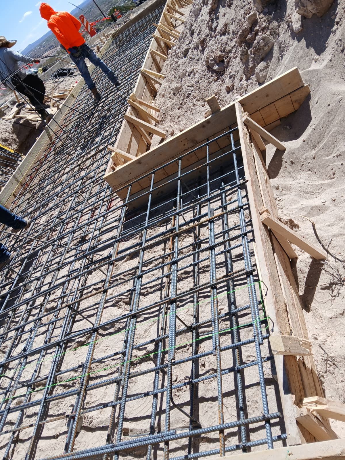 Construction rebar grid and wooden forms in a trench, with workers in orange safety vests nearby