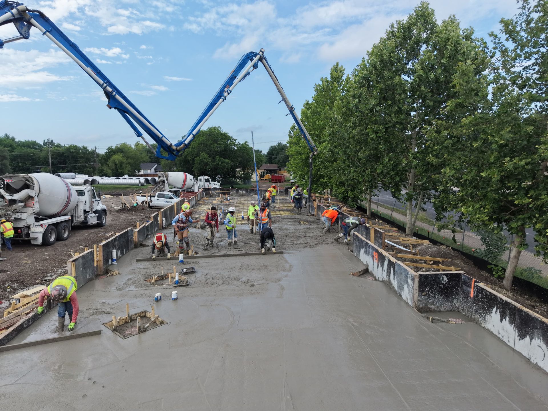 Construction workers pour concrete on a large site with two blue boom pumps and trees in the background