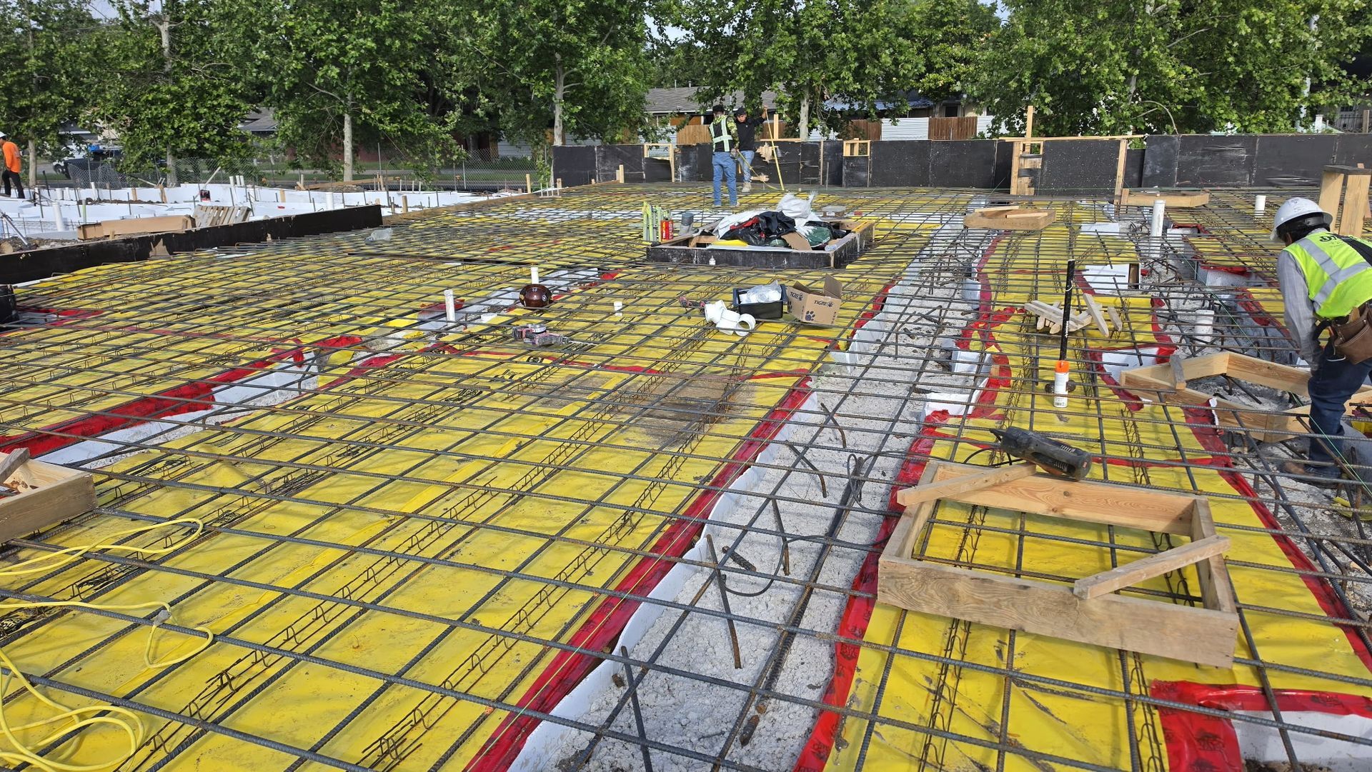 Construction site with rebar grid and yellow forms, workers in safety vests under trees