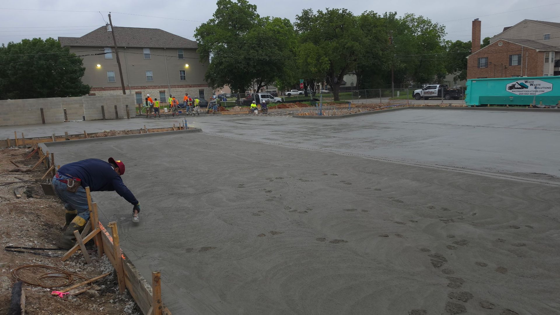 Construction worker smoothing wet concrete in a large outdoor lot near houses and trees