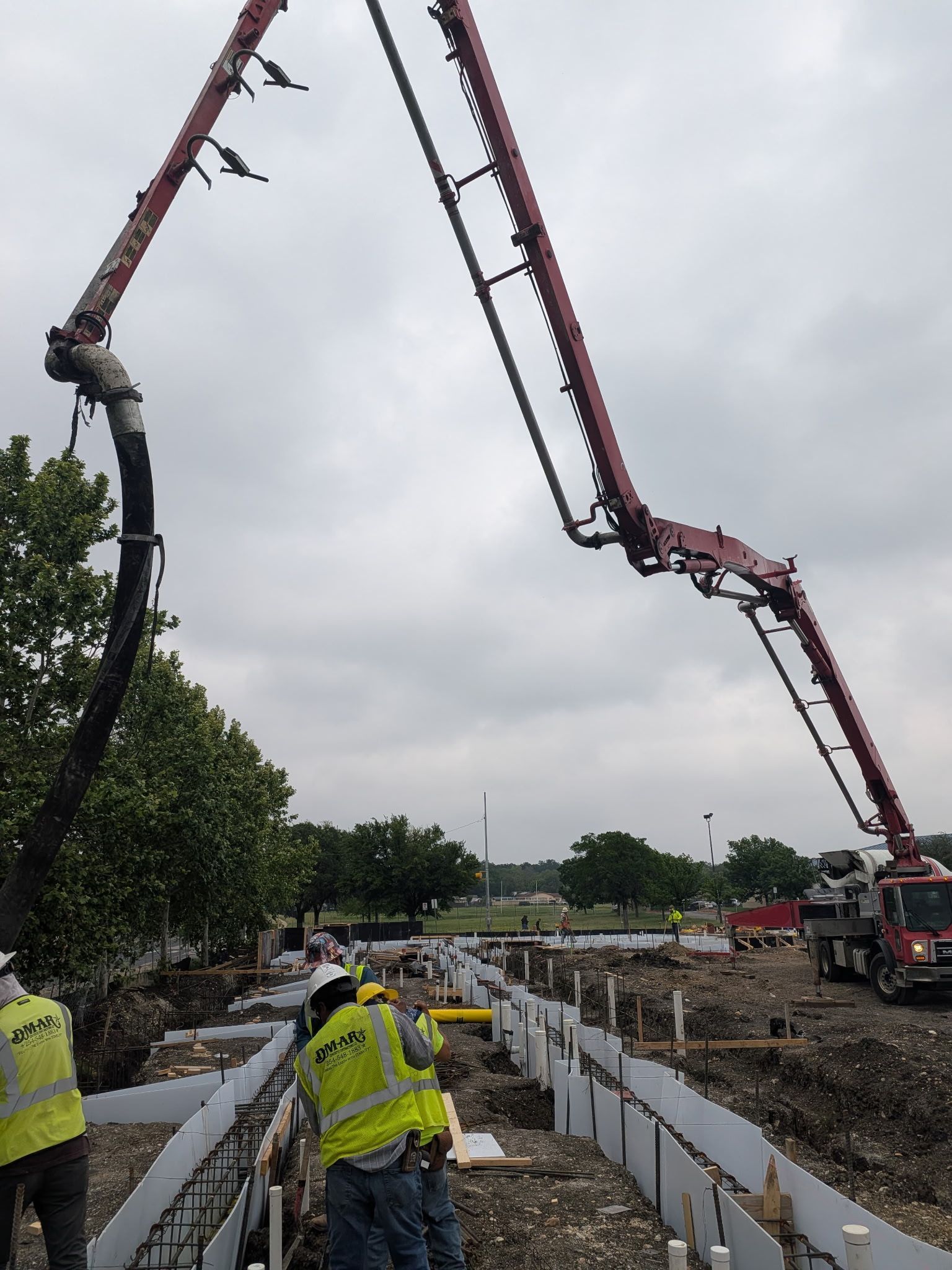 Workers pour concrete from a long boom pump at a construction site under a cloudy sky.