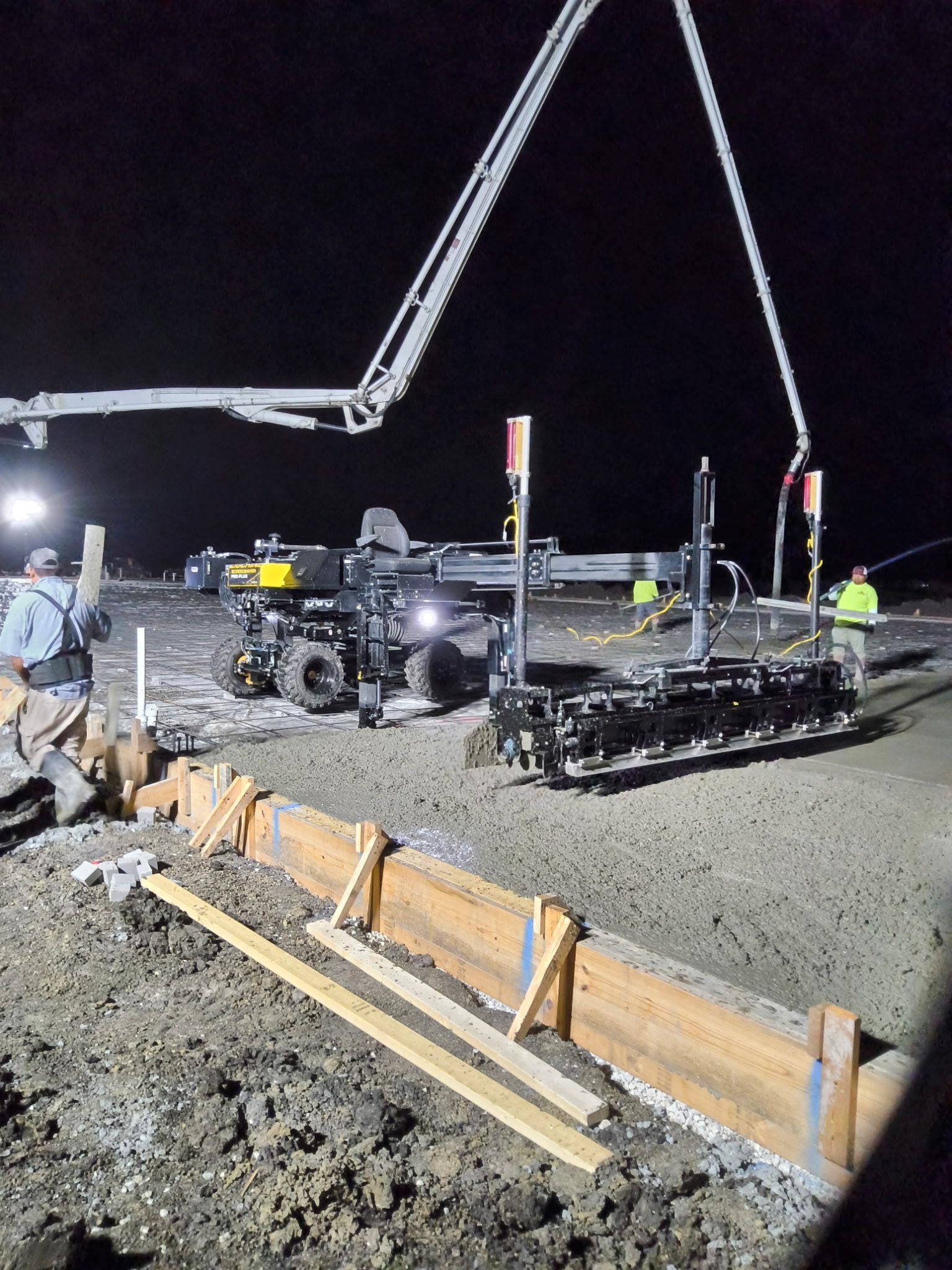 Nighttime road construction with a crane, workers, and equipment pouring concrete into a trench.