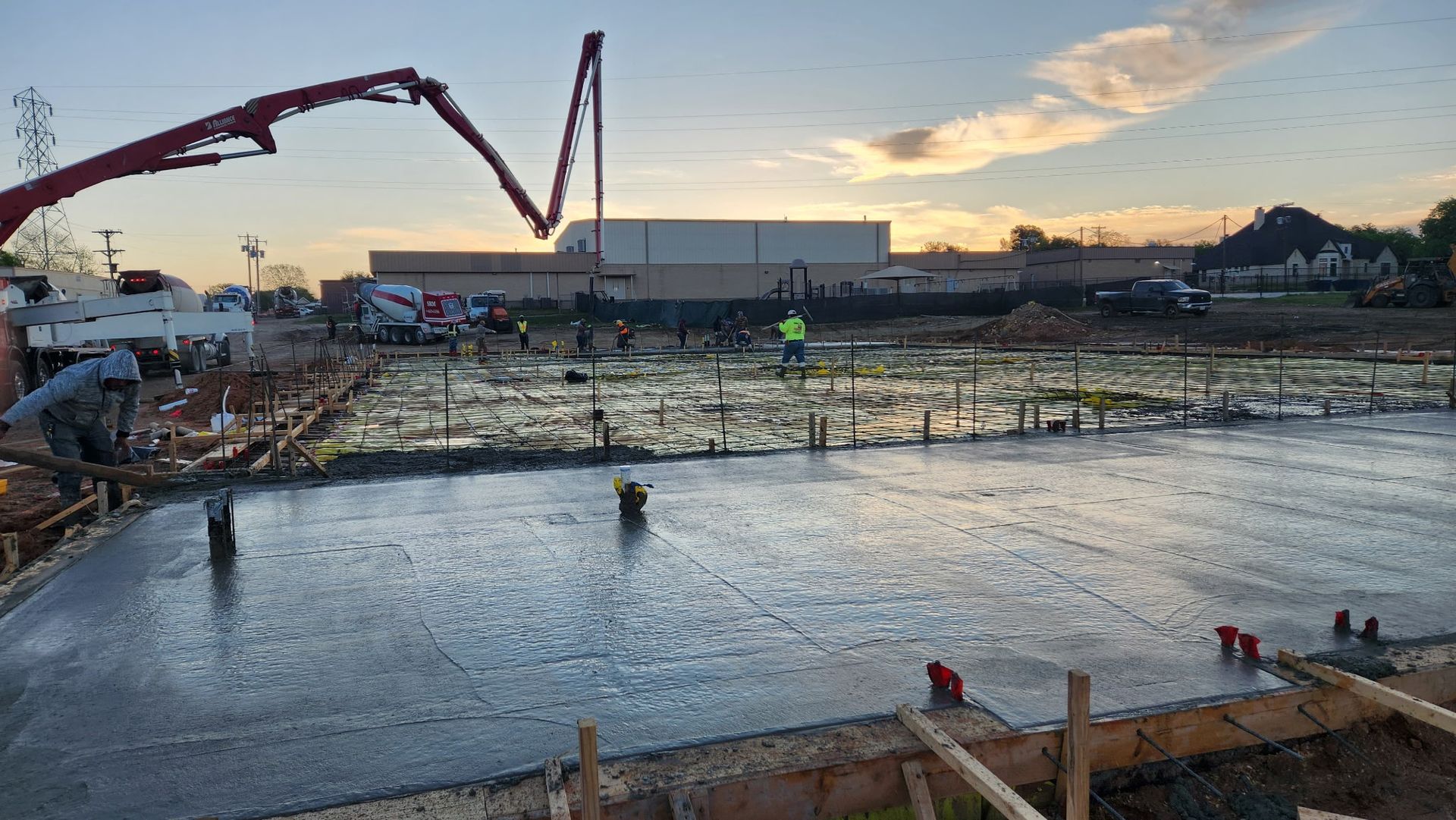 Concrete pump pours into a pond-like construction site at sunset, with workers and buildings in the background