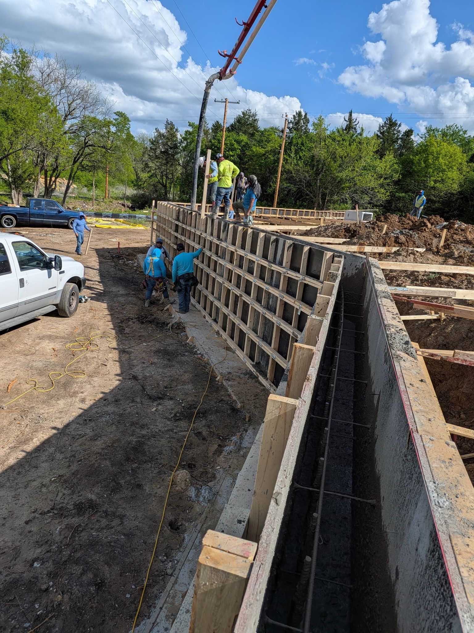 Construction site with workers building a concrete wall beside a trench and parked vehicles under a blue sky
