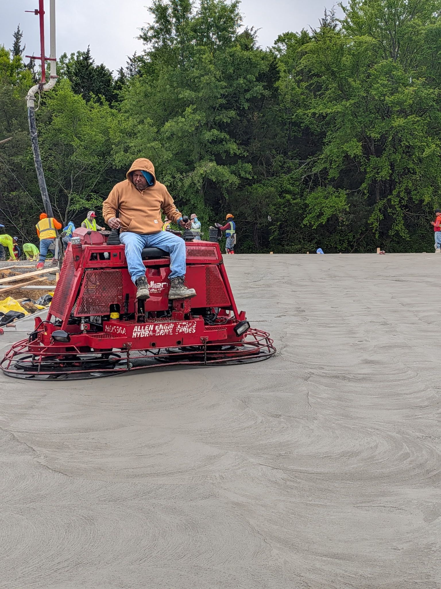 Person riding a red lawn mower on a paved area near trees and flowers
