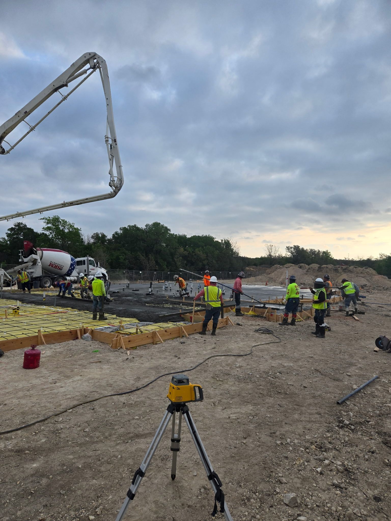 Construction workers pour concrete on a large site at dusk, with a tripod survey camera in the foreground.