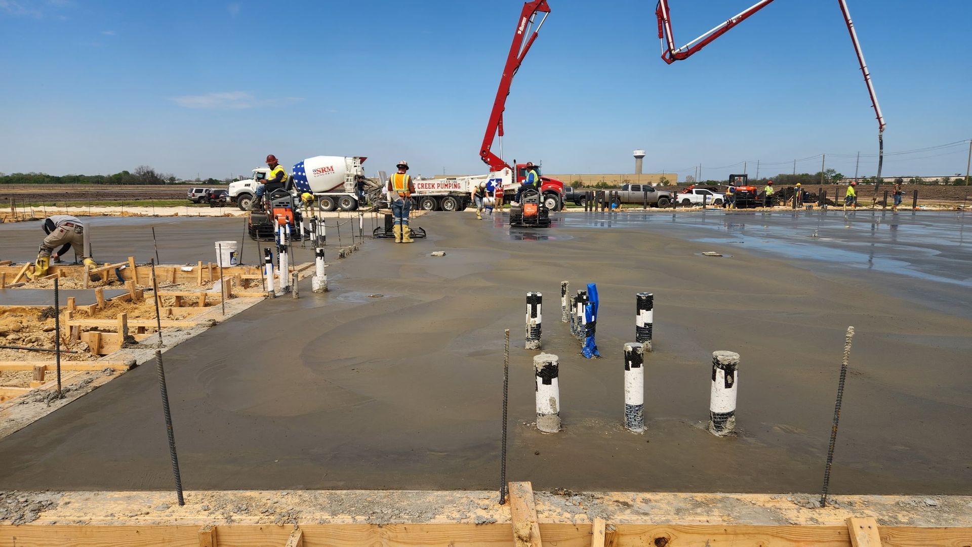 Construction workers pour concrete at a road intersection, with a red pump truck and wet pavement visible.