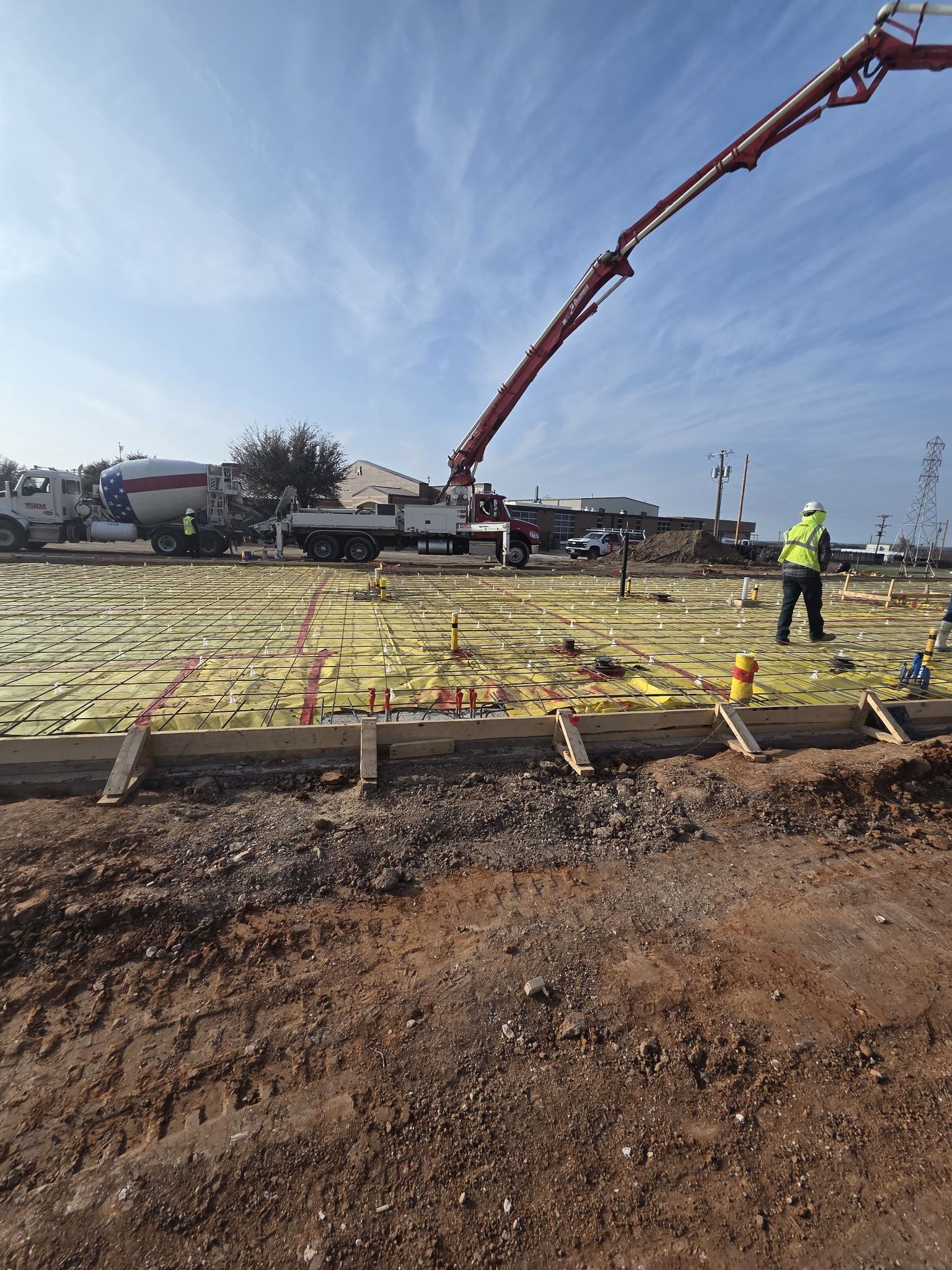 Construction site with concrete pump arm over foundation, worker in high-vis vest, and houses in background